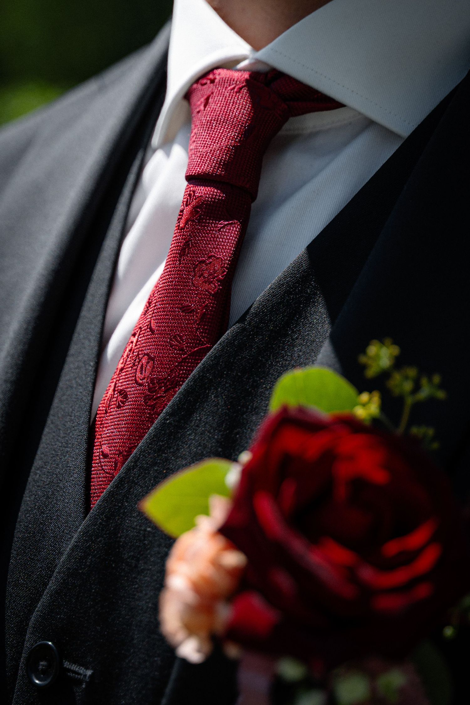 Groom in a dark suit with a red patterned tie and red rose boutonniere on his lapel.