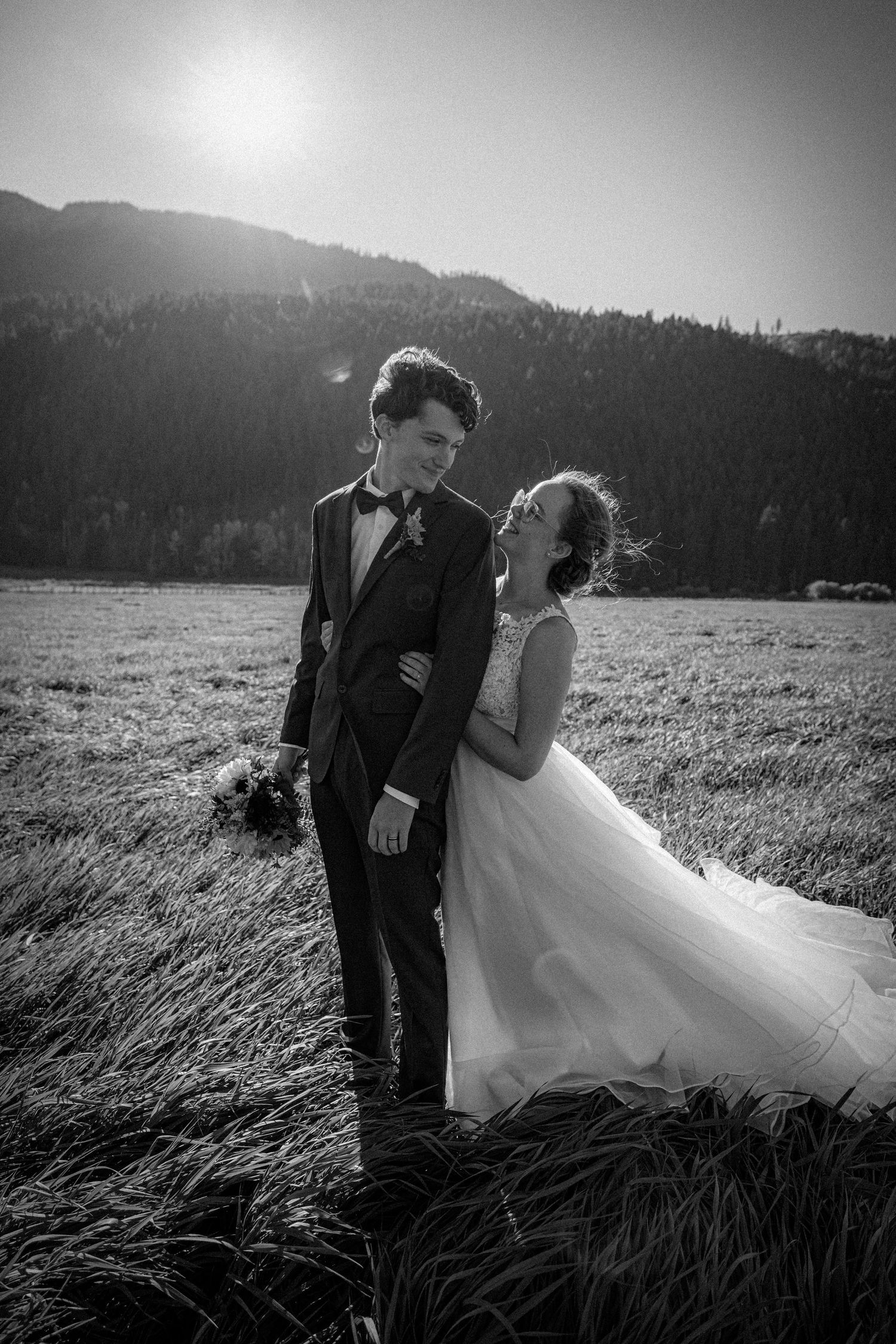 Black and white portrait of laughing bride and groom in sunlit mountain meadow.