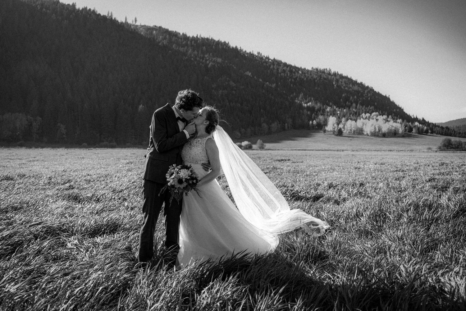 Black and white photo of couple kissing in windswept field with mountain backdrop.