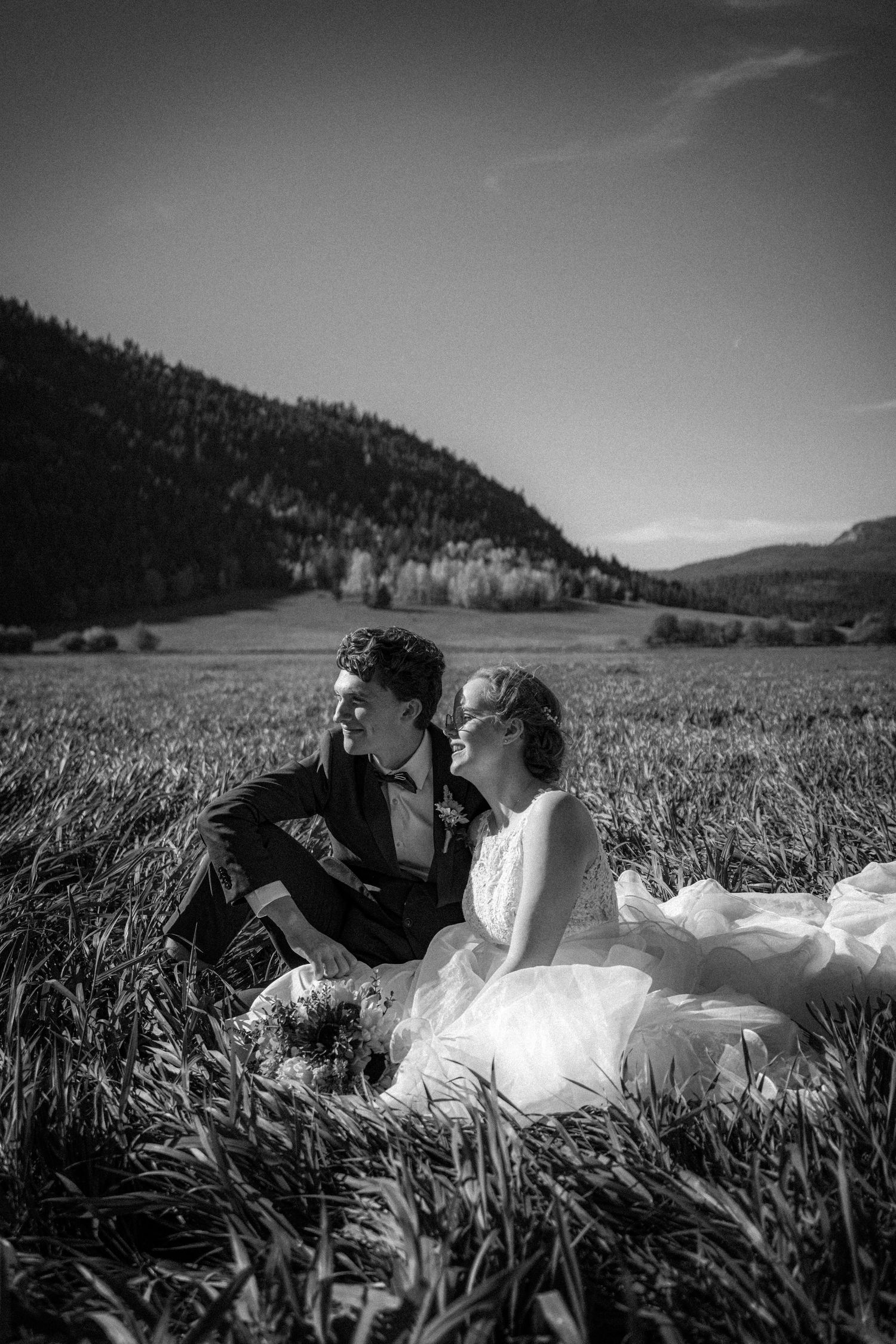 Black and white photo of bride and groom sitting together in a field with mountain backdrop.