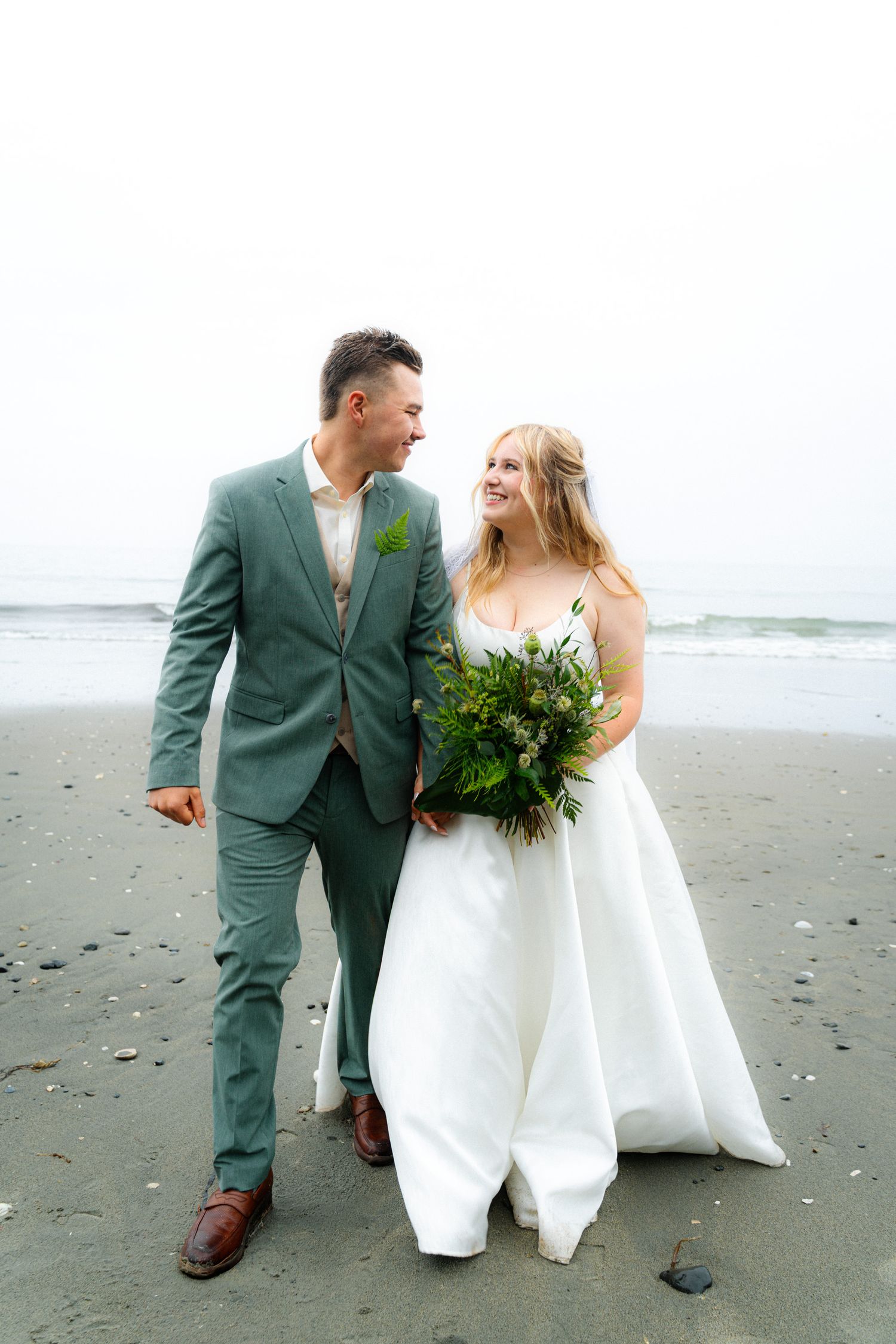 Bride and groom walking on beach, smiling at each other.