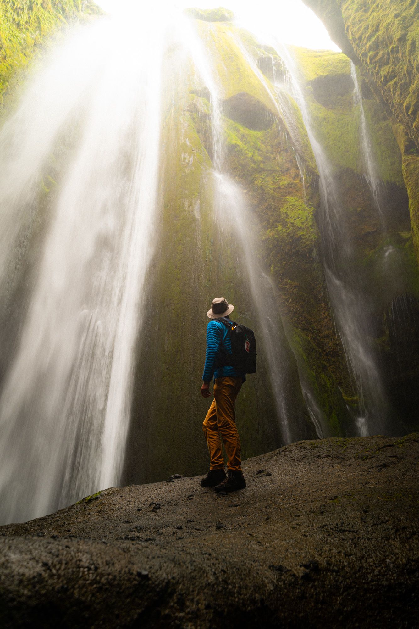 Hiker standing in awe beneath a dramatic cascading waterfall in a mossy canyon.
