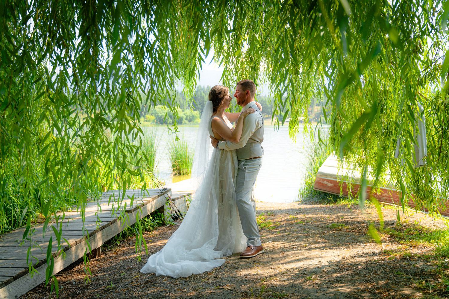 Wedding couple sharing romantic kiss under weeping willow branches near scenic lakeside dock.