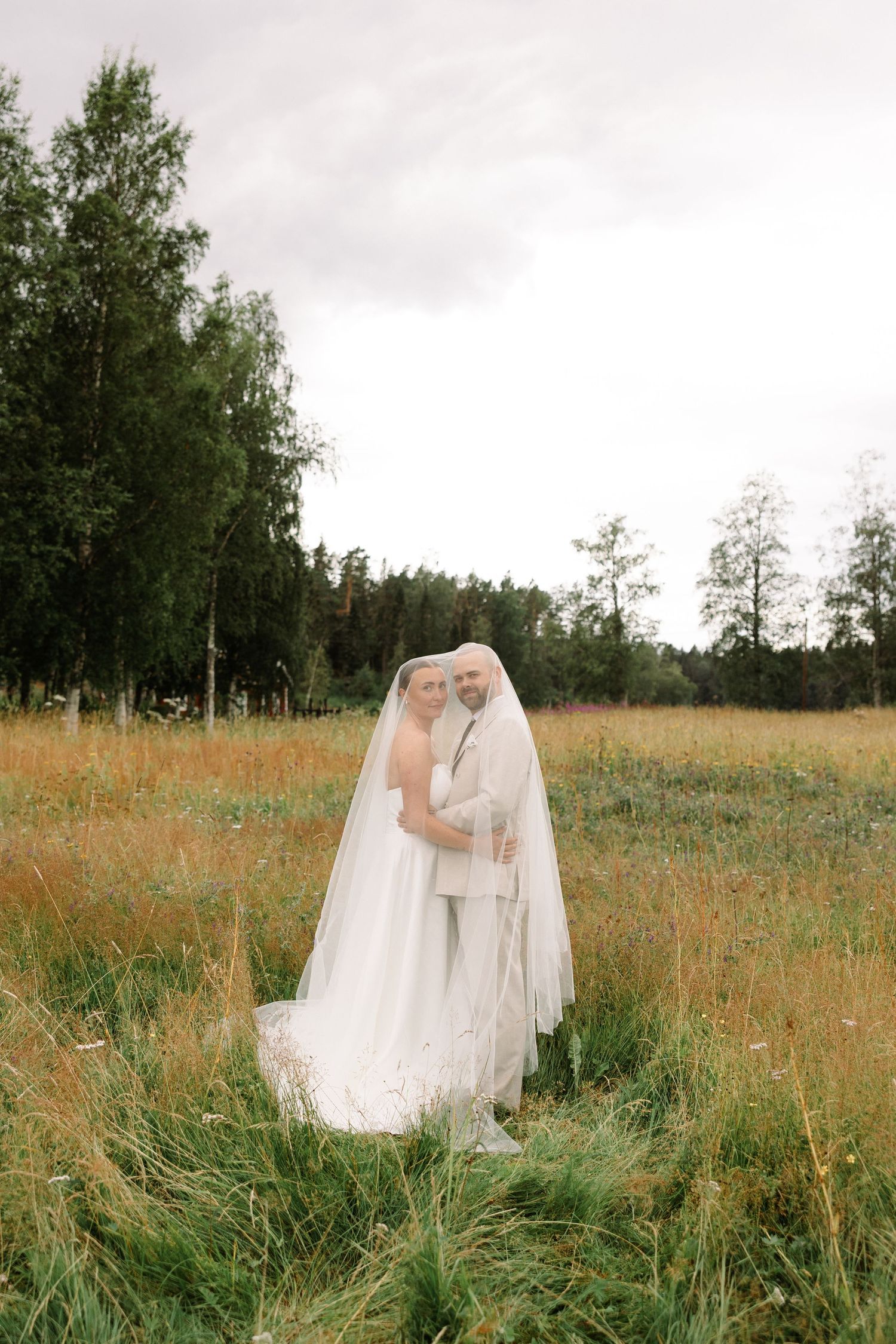 Bride and groom embracing under a flowing white veil in a golden meadow with lush green trees in the background.