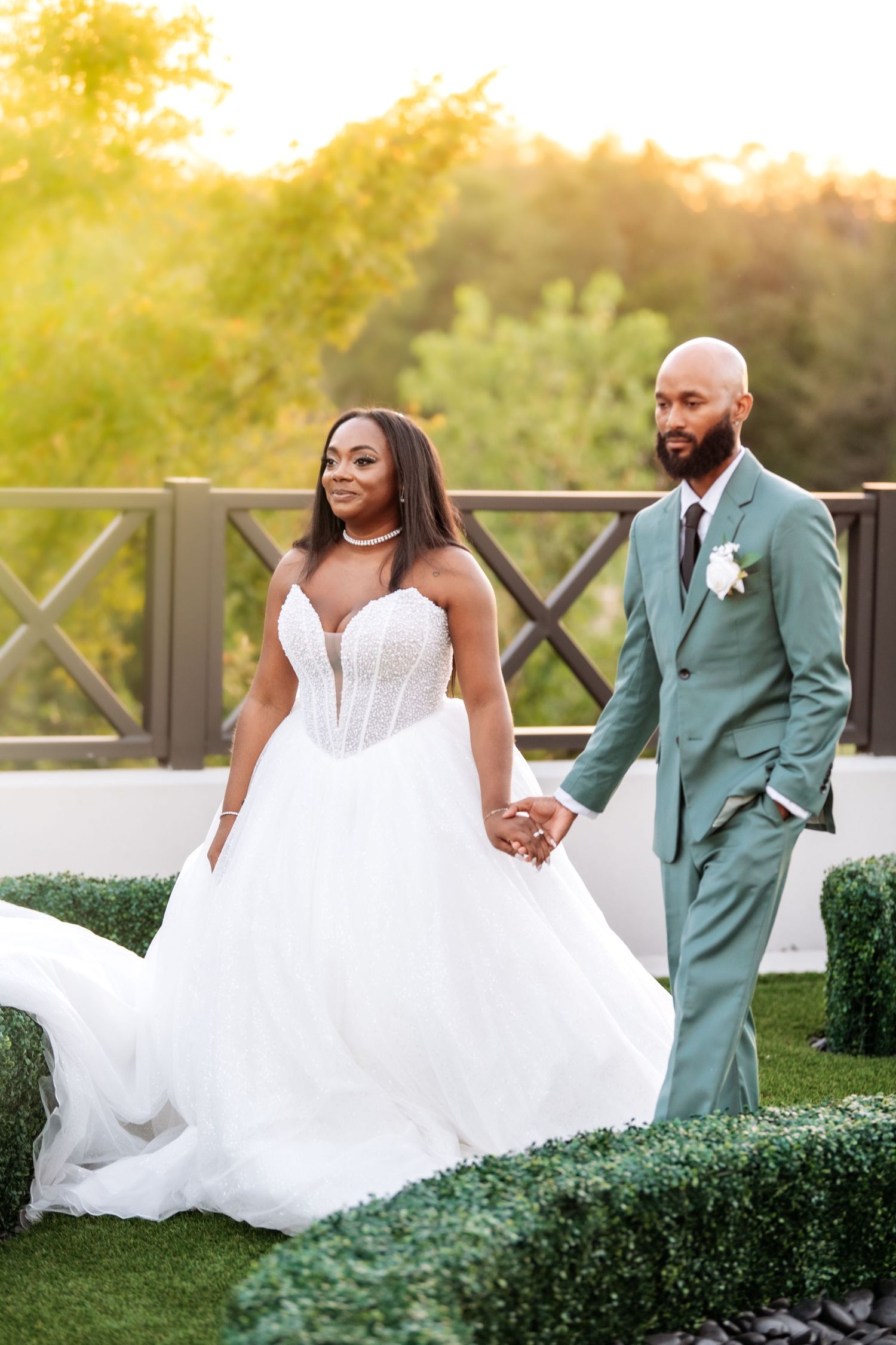 Bride in white ballgown and groom in sage green suit hold hands outdoors at golden hour wedding ceremony.