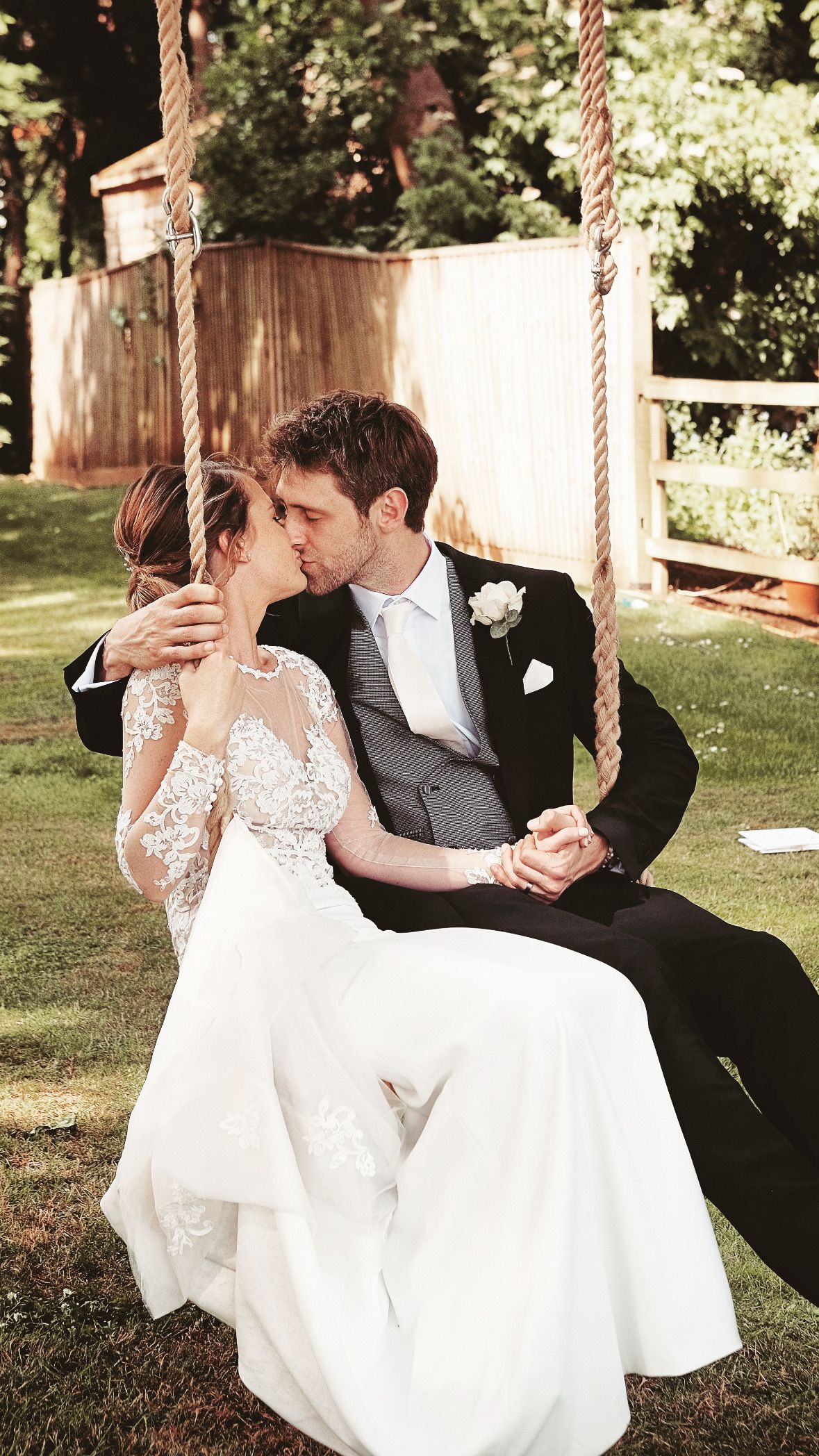 Romantic couple shares a tender kiss while sitting together on a wooden swing in a sunlit garden setting.