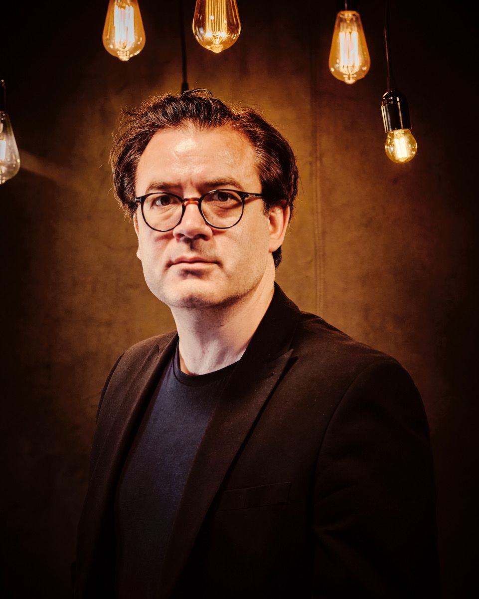 Professional headshot with vintage Edison bulbs hanging in background against dark brown wall.