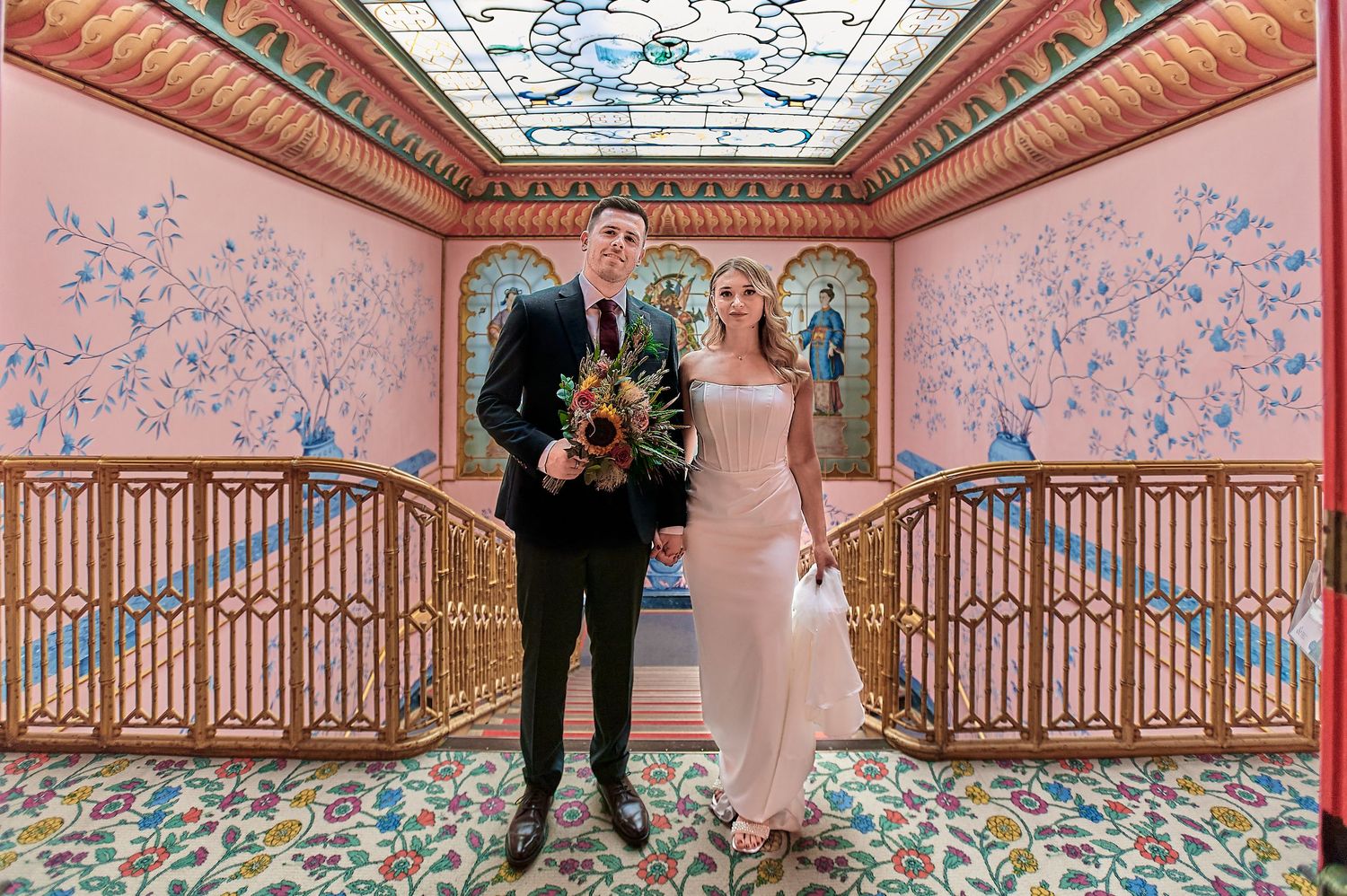 Bride and groom stand on ornate staircase with stained glass ceiling and colorful floral walls in elegant wedding venue.