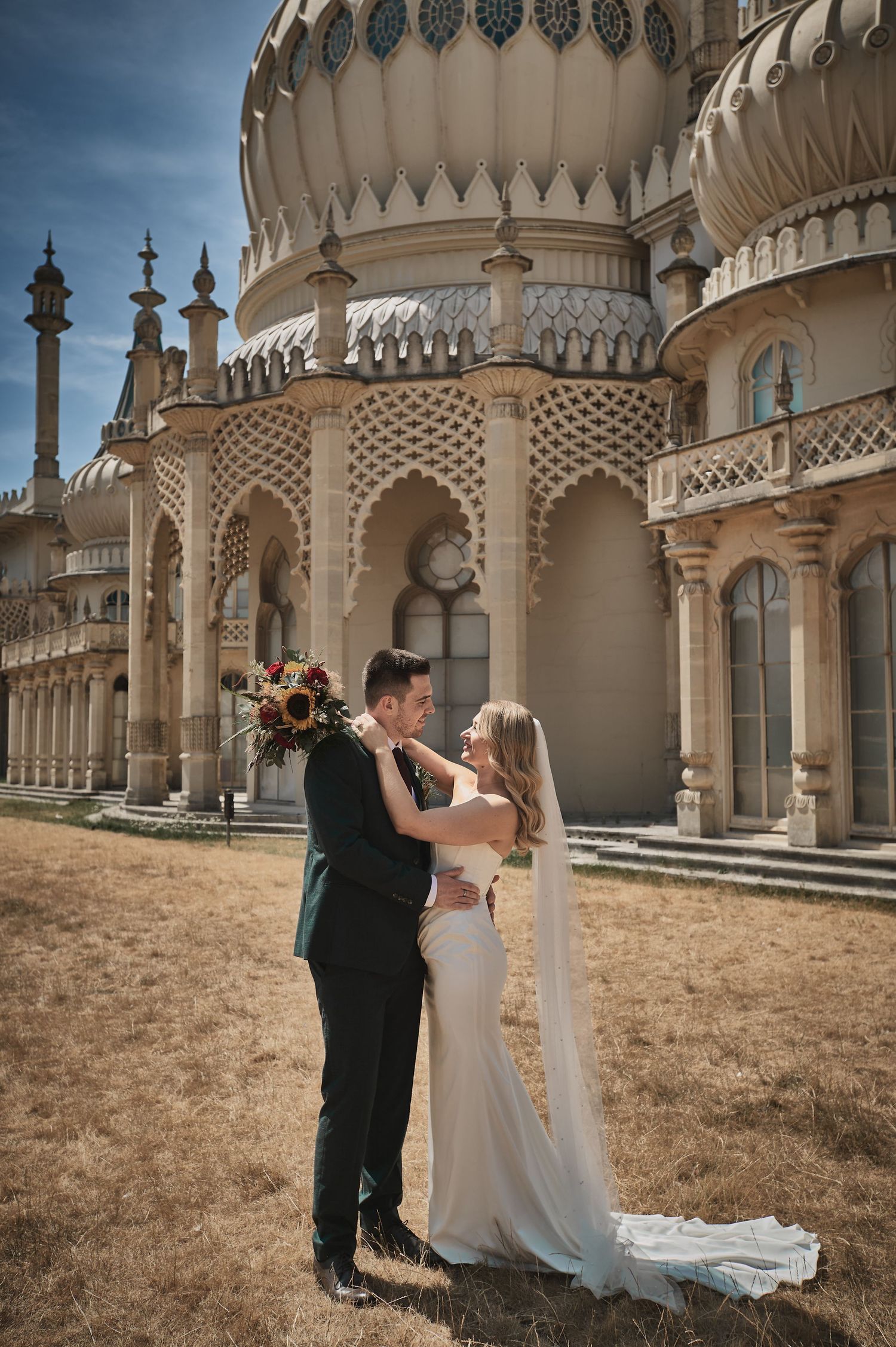 Bride and groom embrace in front of Brighton Royal Pavilion, bride holding bouquet in elegant white gown with veil.