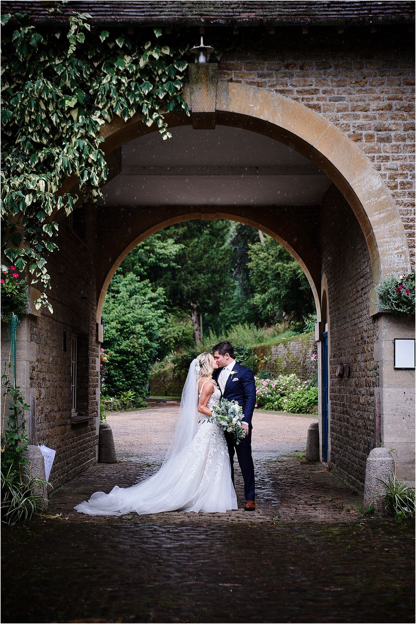 A Surrey couple shares a tender moment under a brick archway with ivy growing along the wall while posing for their wedding photo.