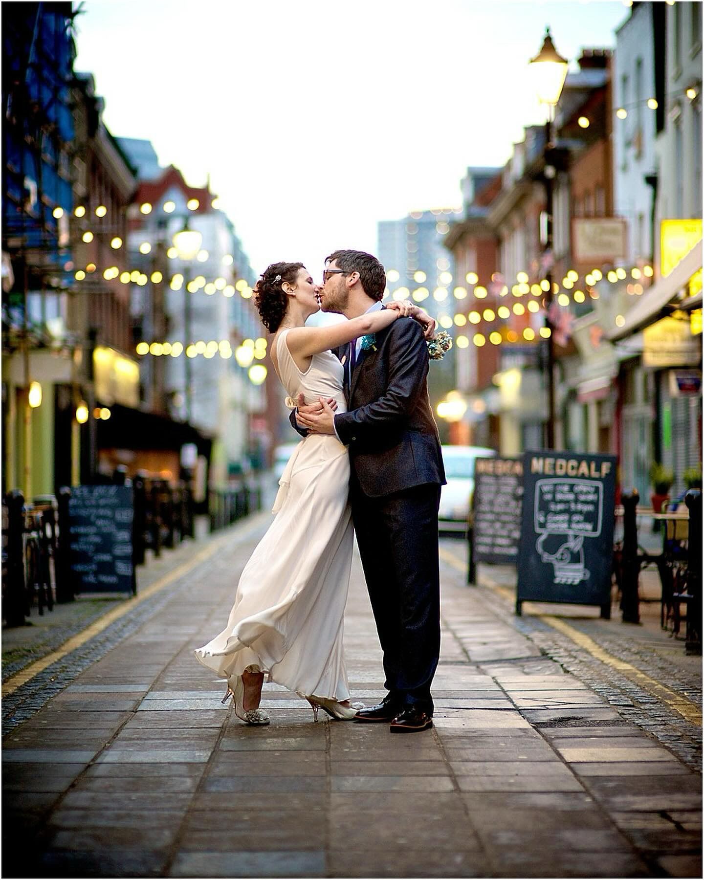 A romantic couple dances and dips on a charming cobblestone street illuminated by overhead string lights at dusk.