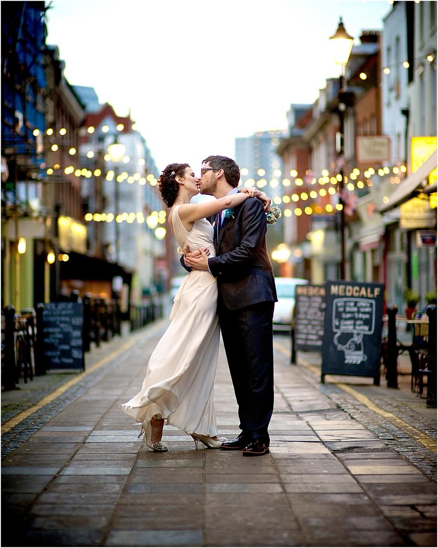 A romantic couple embraces and dips on a charming street at dusk with string lights glowing overhead.