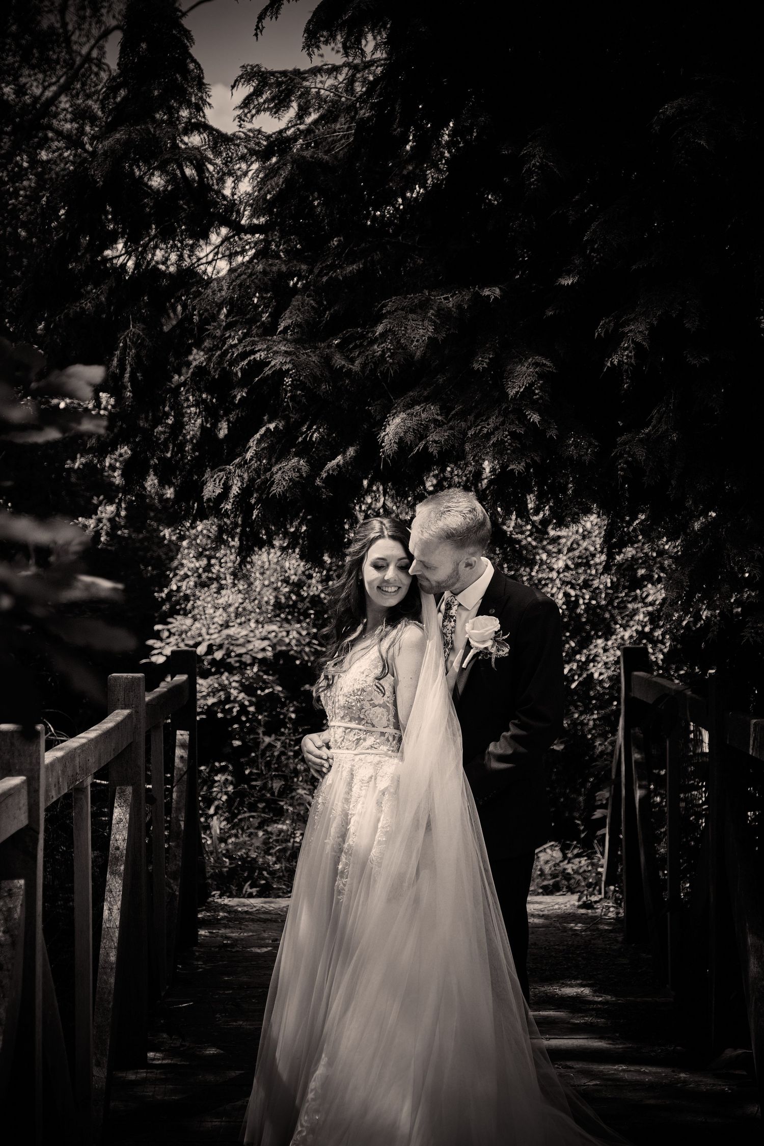 A romantic black and white wedding photo of a couple embracing on a wooden bridge surrounded by lush trees.