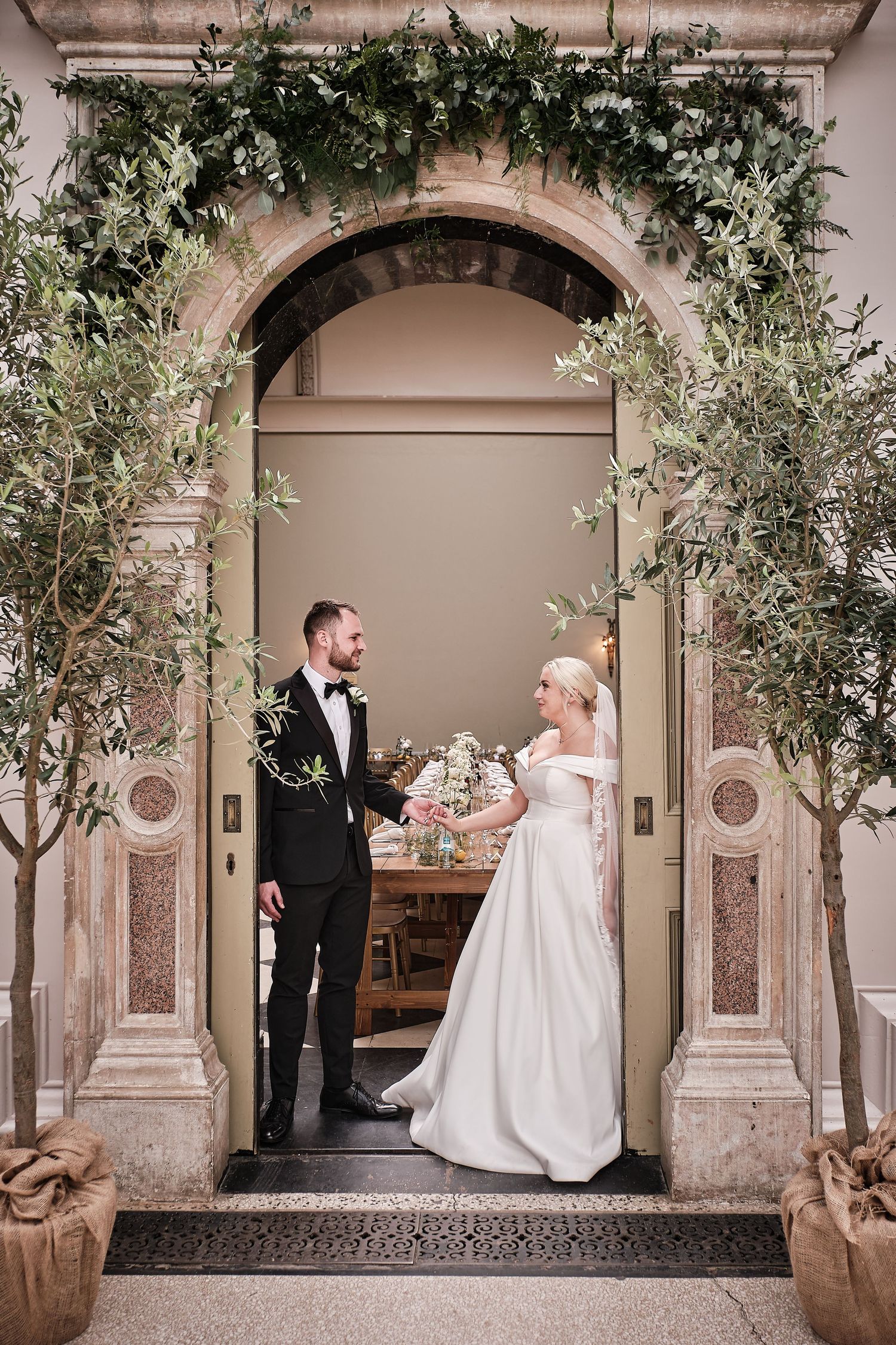 Elegant wedding couple stands in ornate stone archway decorated with greenery and olive branches at Hampton Court House entrance.