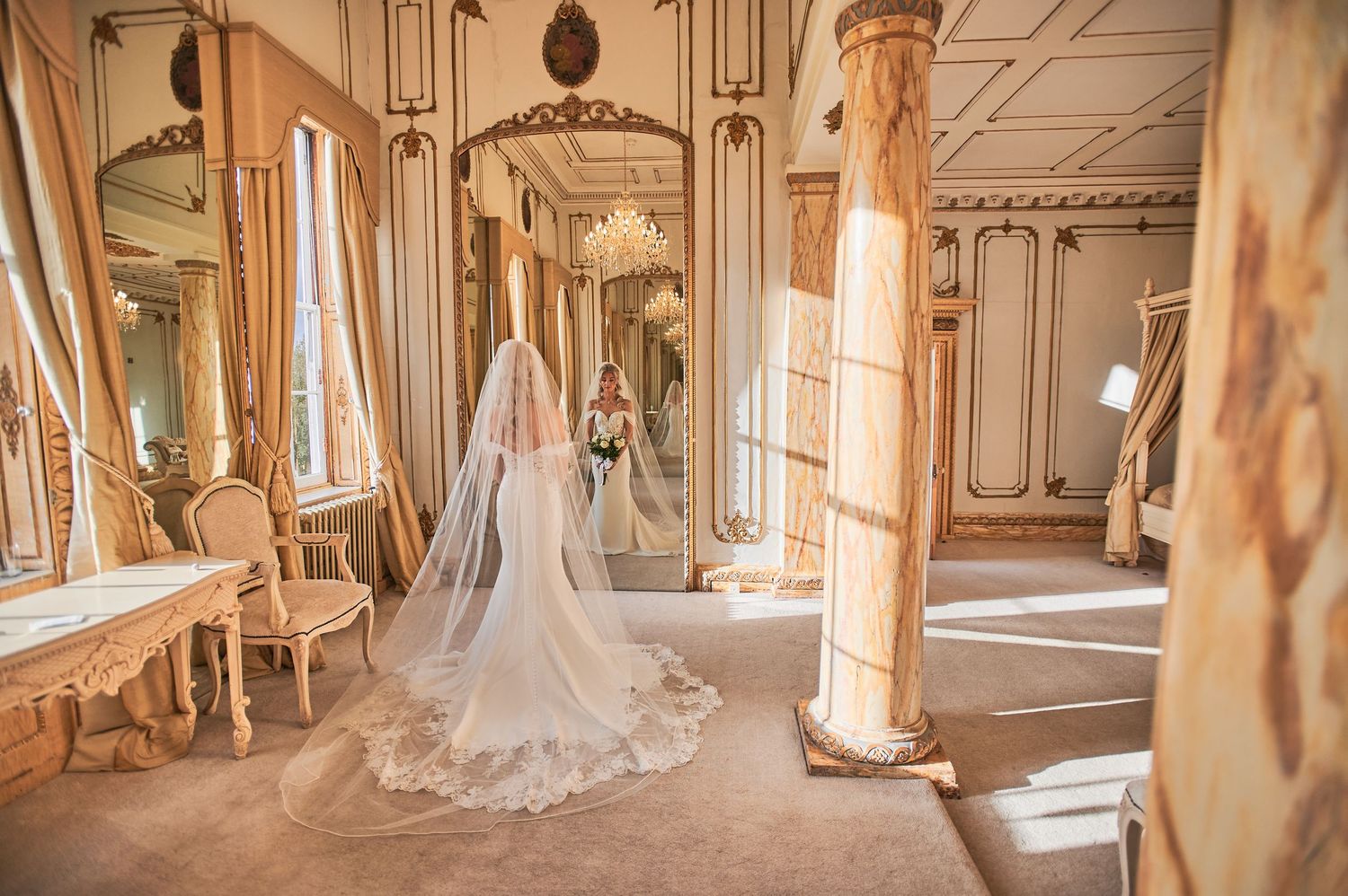 Bride in flowing white gown and long veil stands in ornate golden ballroom with tall mirrors and marble columns.