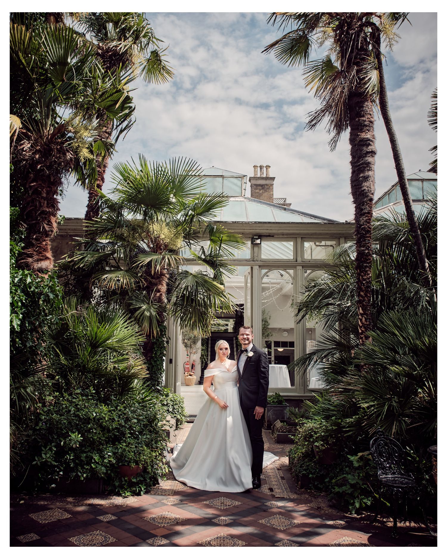 Couple poses together in a lush garden courtyard with palm trees and white architecture in the background.