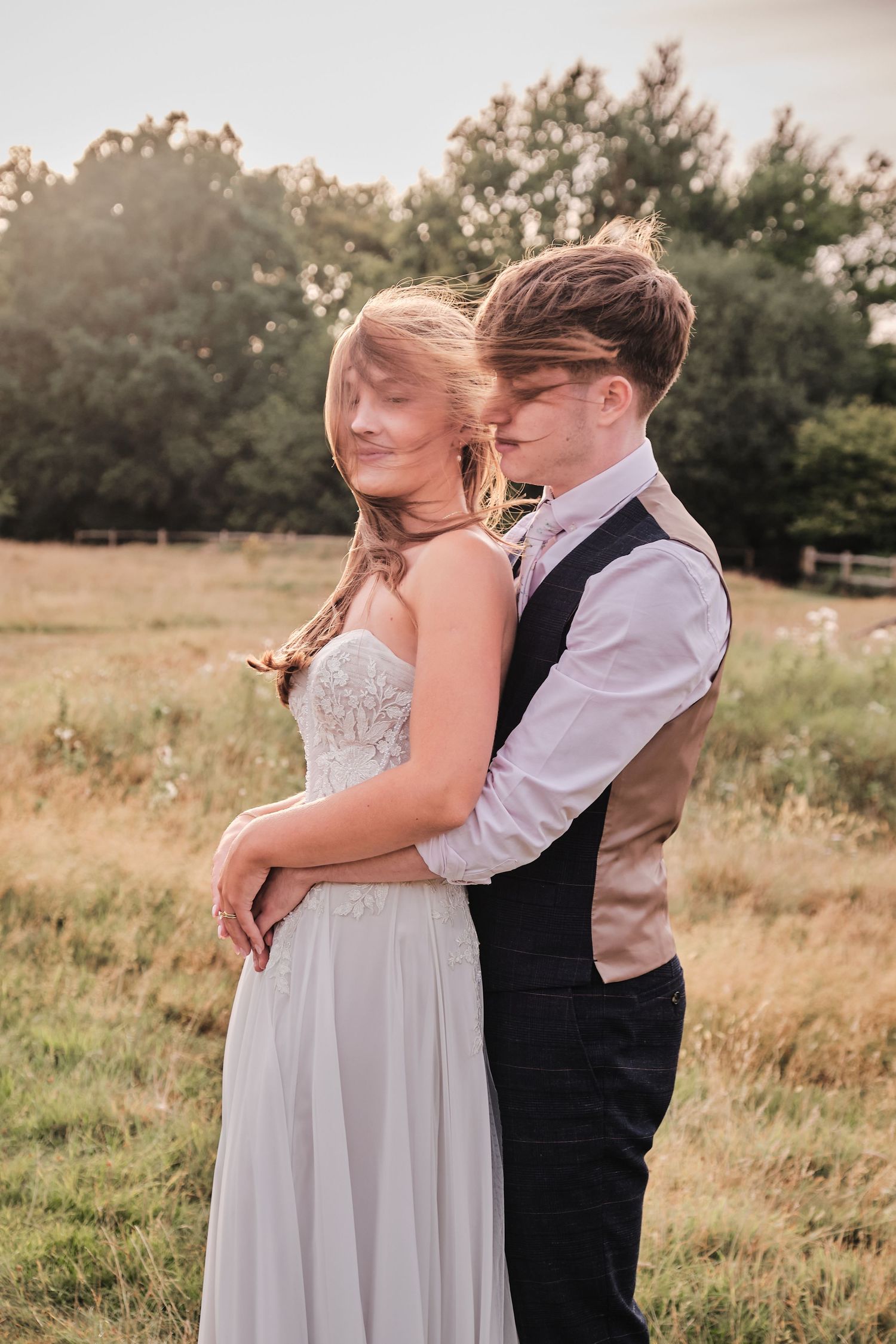 A couple in formal attire embraces tenderly in a sunlit meadow during a romantic outdoor photo session.