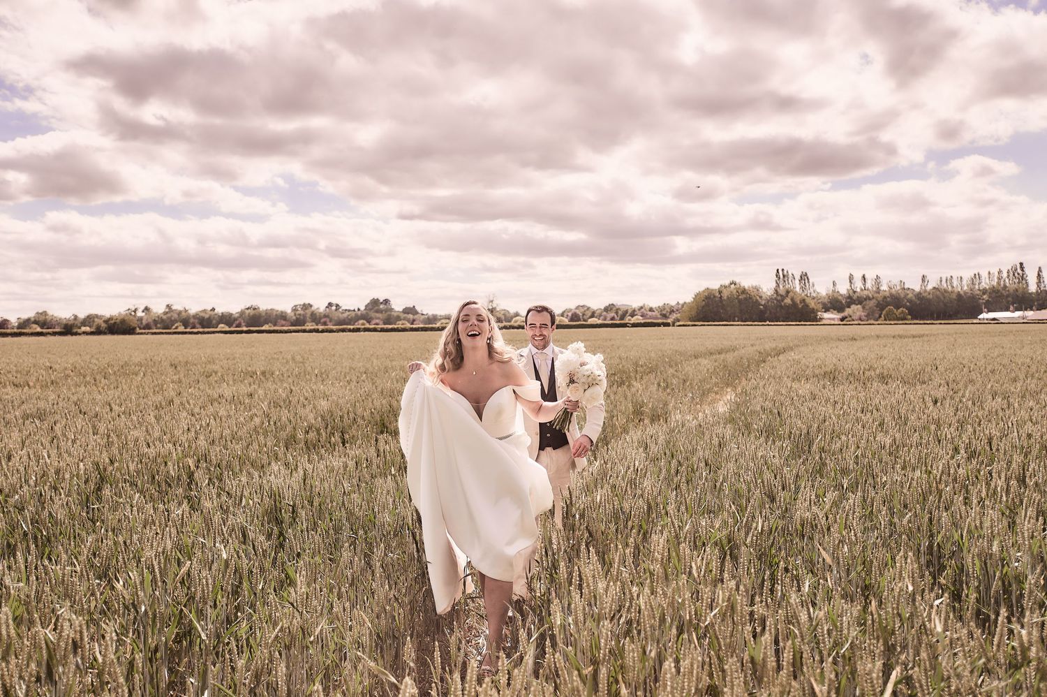 Bride and groom running through a lush green wheat field under a dramatic cloudy sky on their wedding day.