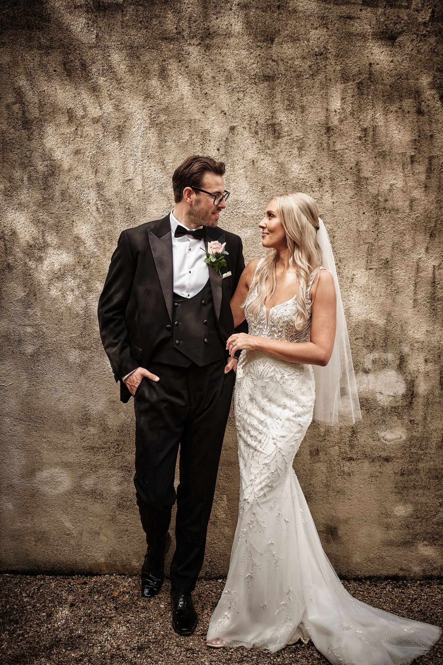 Classic wedding portrait of a bride in a lace gown and groom in tuxedo against textured brown wall. A surrey wedding photographer