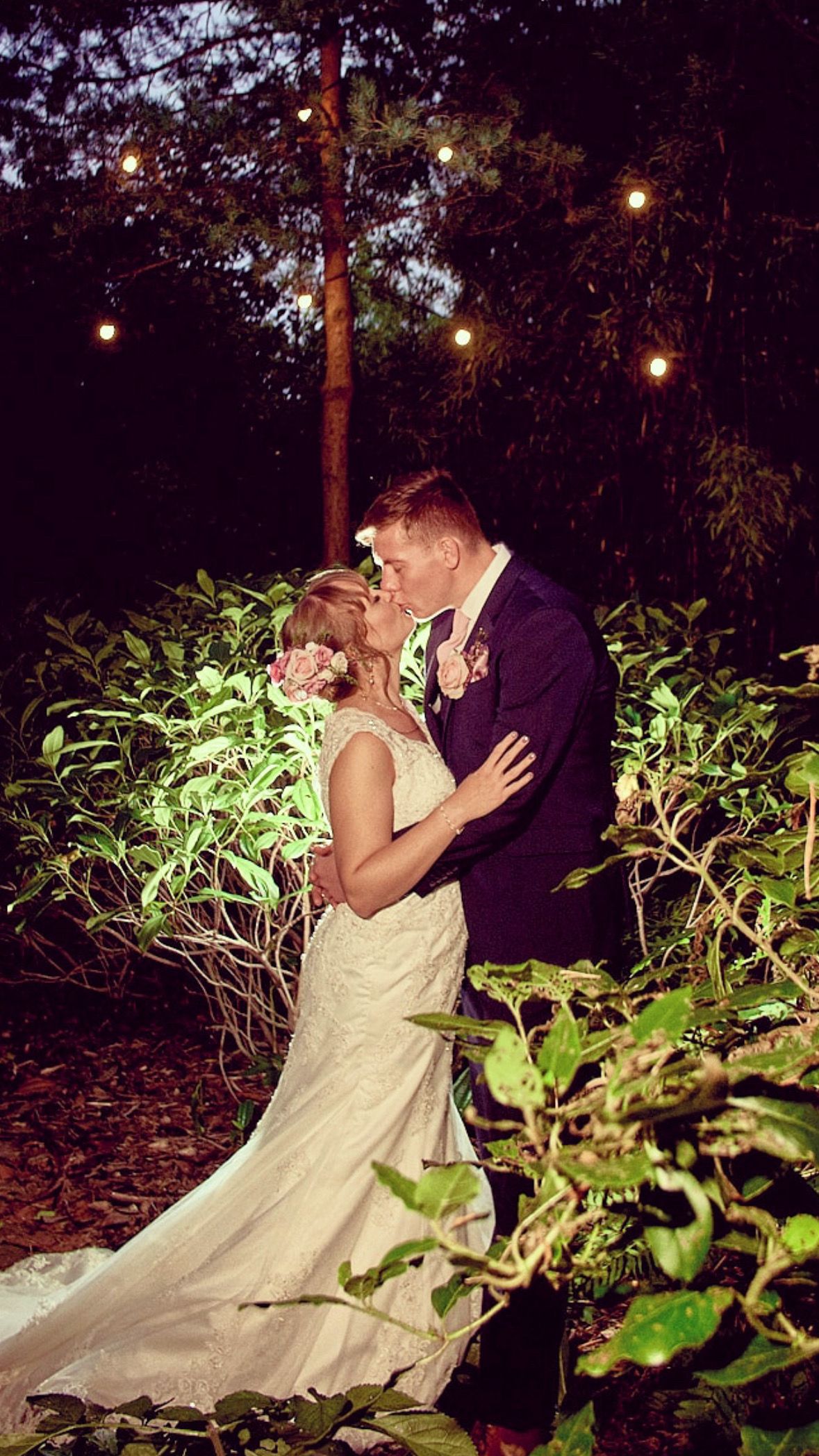 A romantic couple shares a kiss in a garden at night with string lights glowing overhead in a wedding setting.