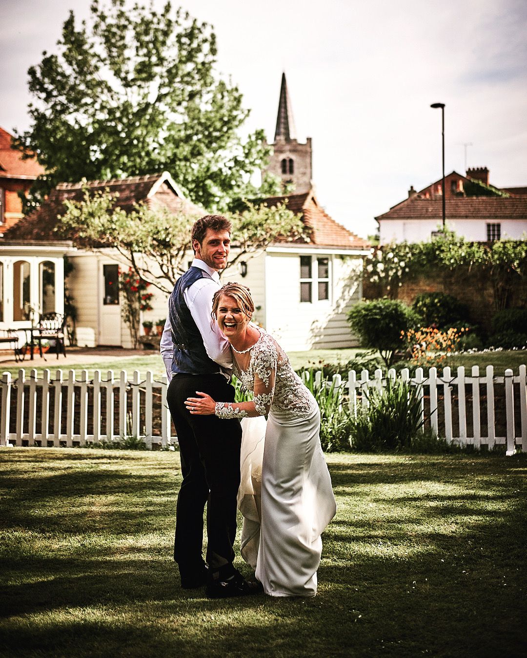 A romantic wedding photo taken in a garden setting with a white picket fence and church steeple visible in the background.