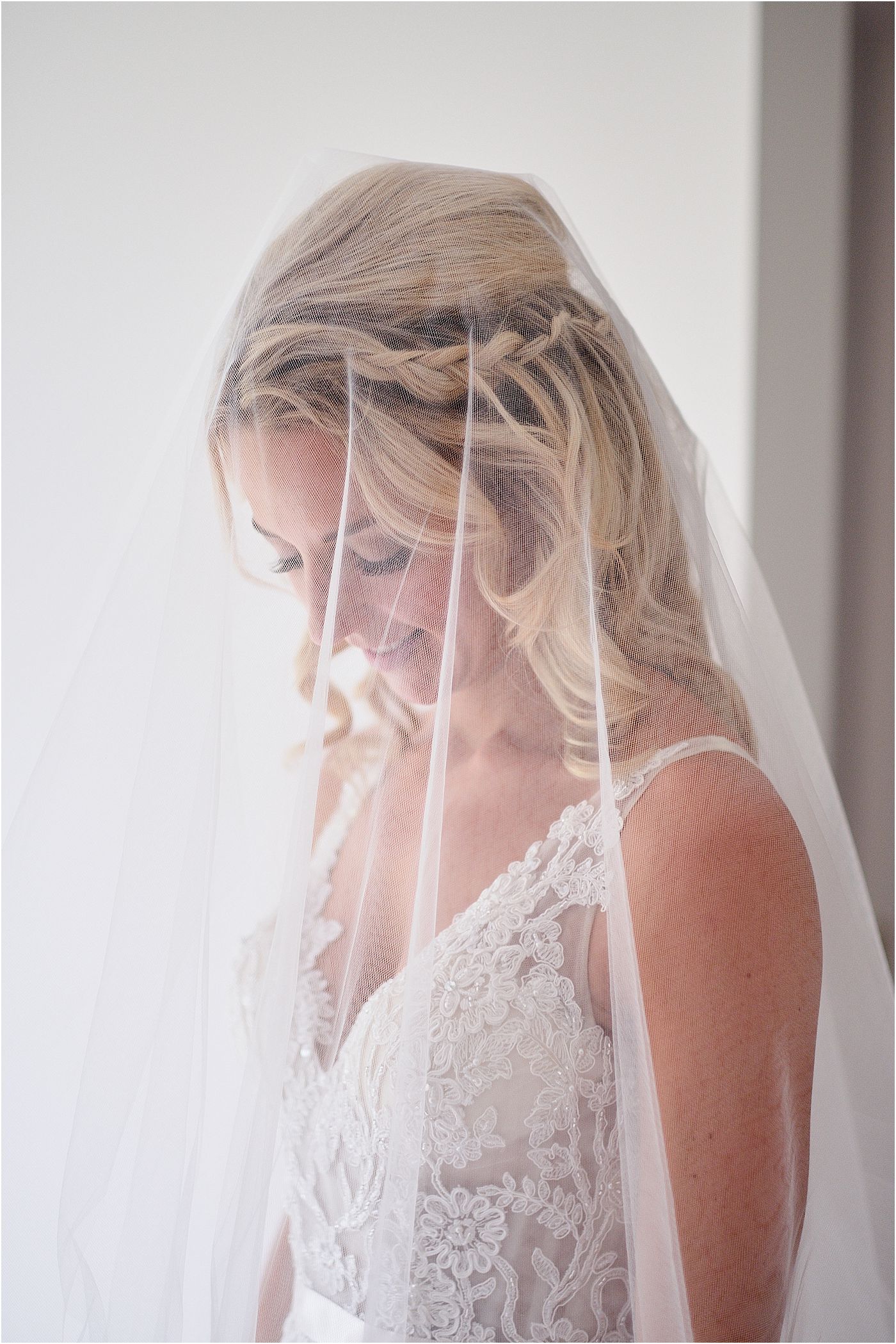 A Surrey bride in a delicate lace wedding gown stands near a window with a sheer veil draping down.