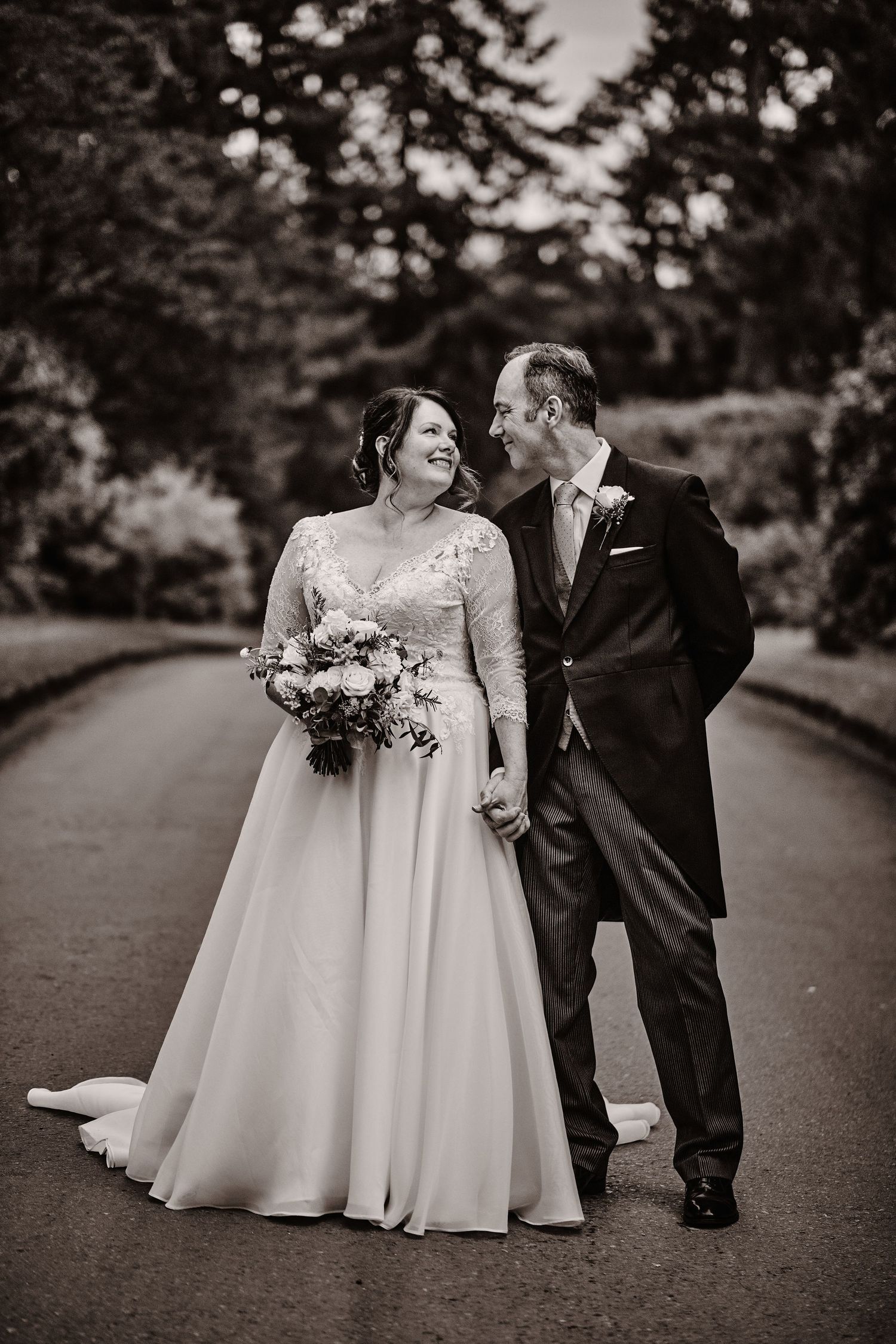 A black and white wedding photo of a couple sharing a joyful moment on a tree-lined path at Ramster Hall  Surrey Wedding PHotographer