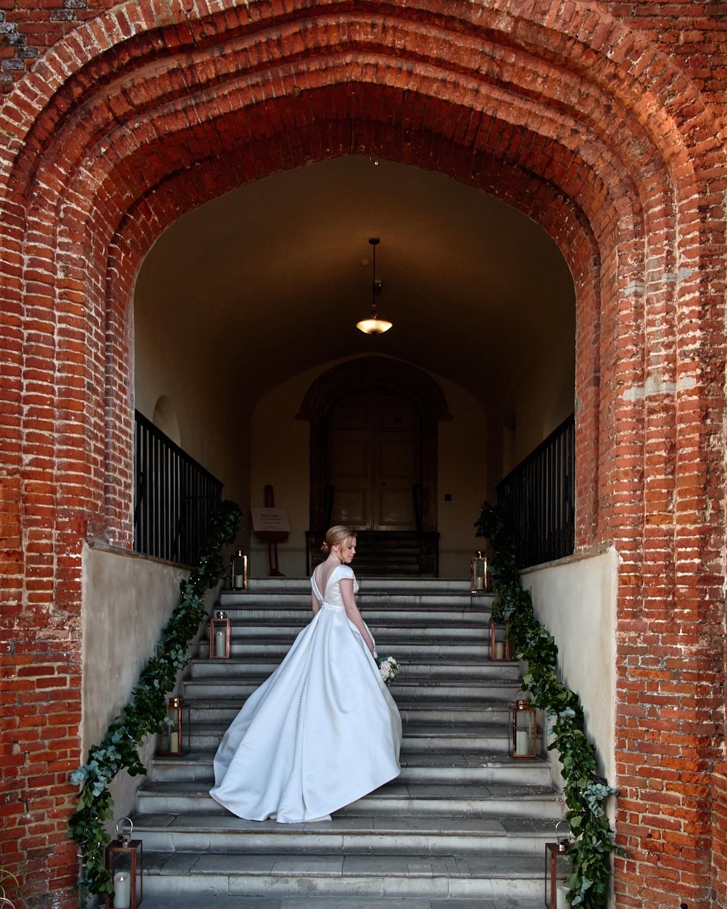 A white wedding dress flows gracefully down brick-arched stairs decorated with greenery garlands and an overhead light.