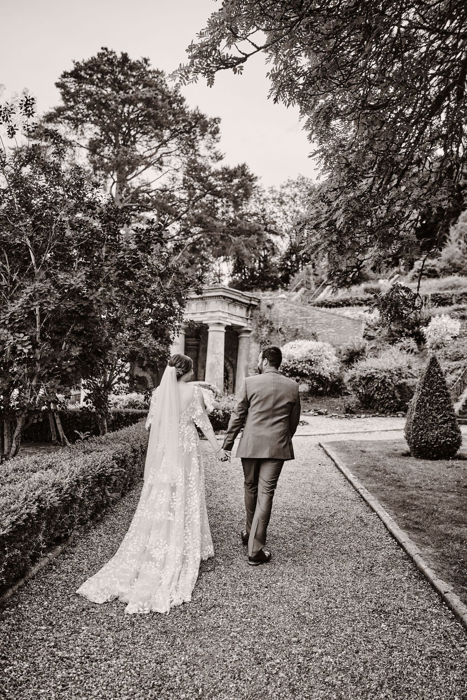 A couple walks along a garden path towards a stone structure in a romantic black and white wedding photo.