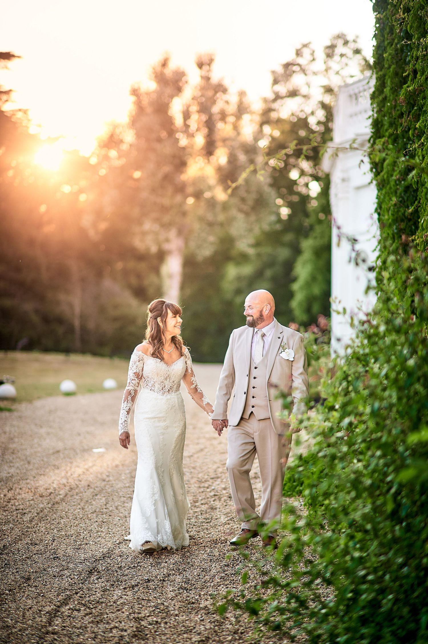 Bride and groom walk hand-in-hand on a gravel path at golden hour, with lush greenery and warm sunlight behind them.