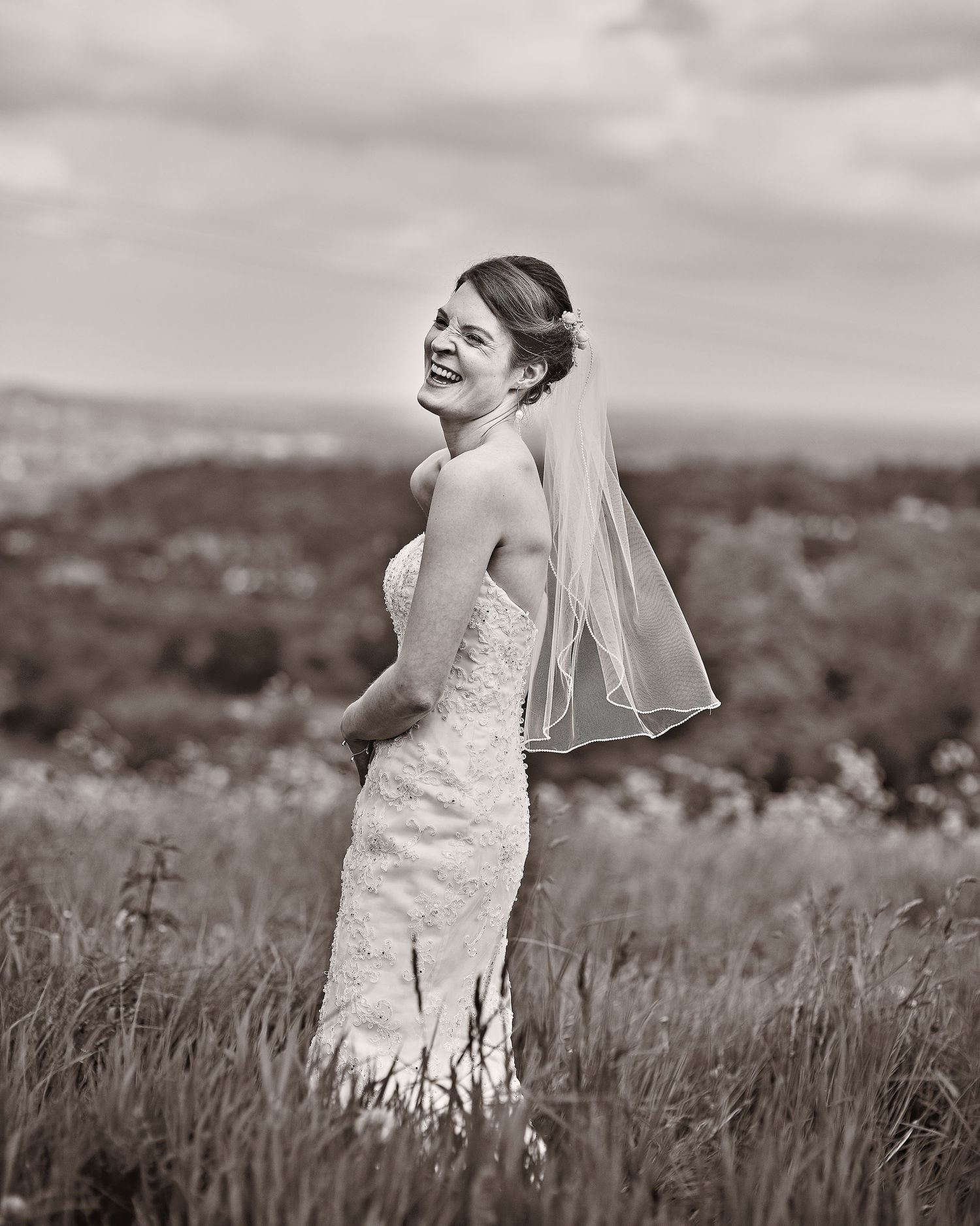 A bride in a lace wedding dress and veil poses outdoors in a field with a scenic landscape backdrop in black and white. Denbies Wine estate