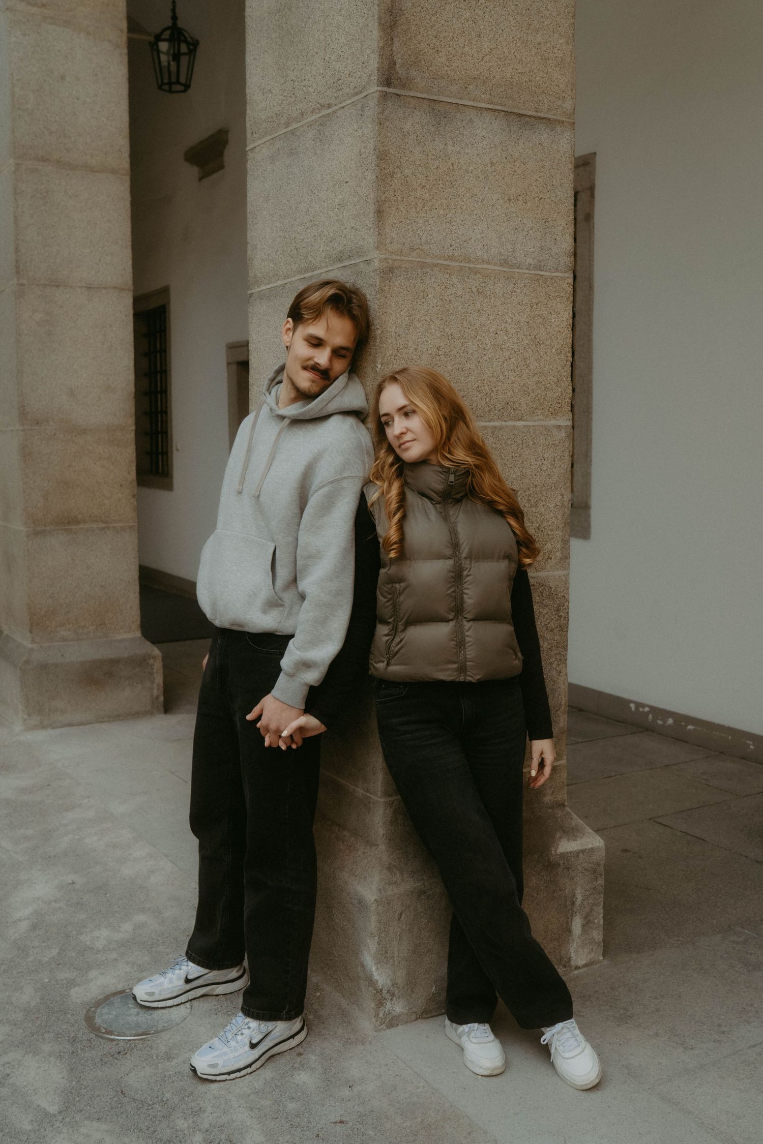 Young couple holding hands leaning against stone pillar, wearing casual streetwear outfits outdoors.
