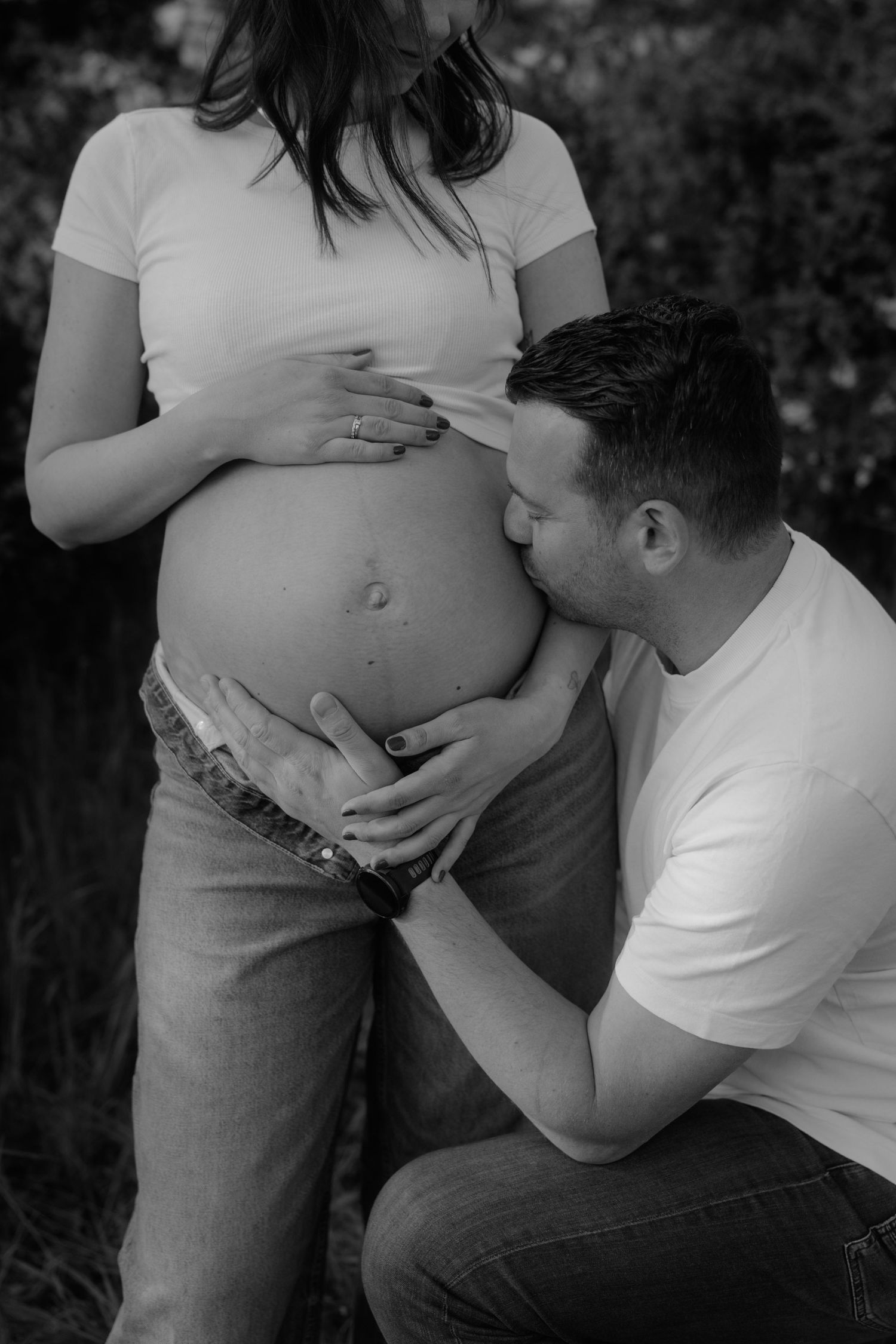 Black and white maternity photo of a man kissing pregnant woman's belly outdoors, both wearing white shirts and jeans.