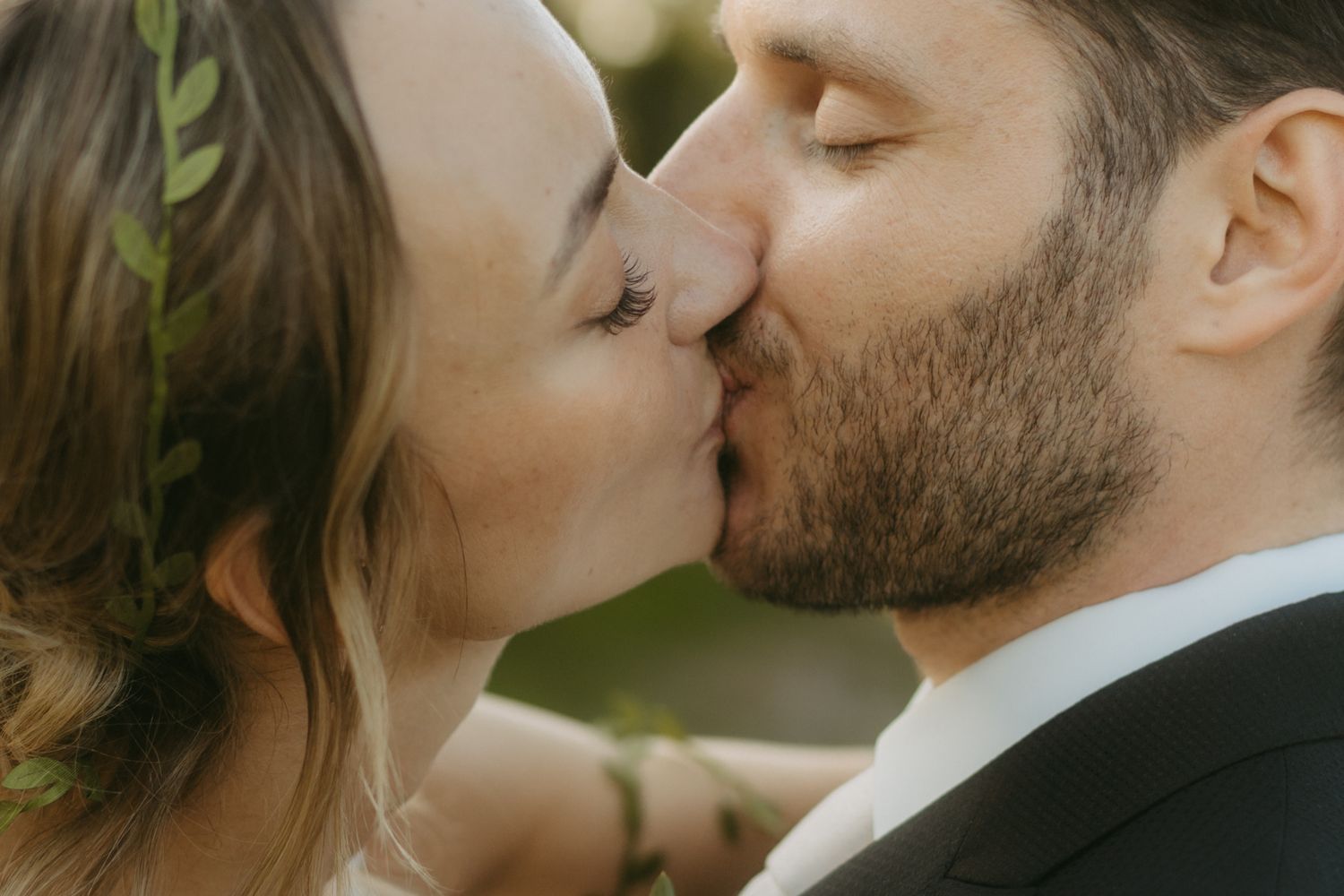 Close-up of a bride with a leafy hair vine and a bearded groom in a dark suit sharing a tender kiss outdoors on their wedding day.
