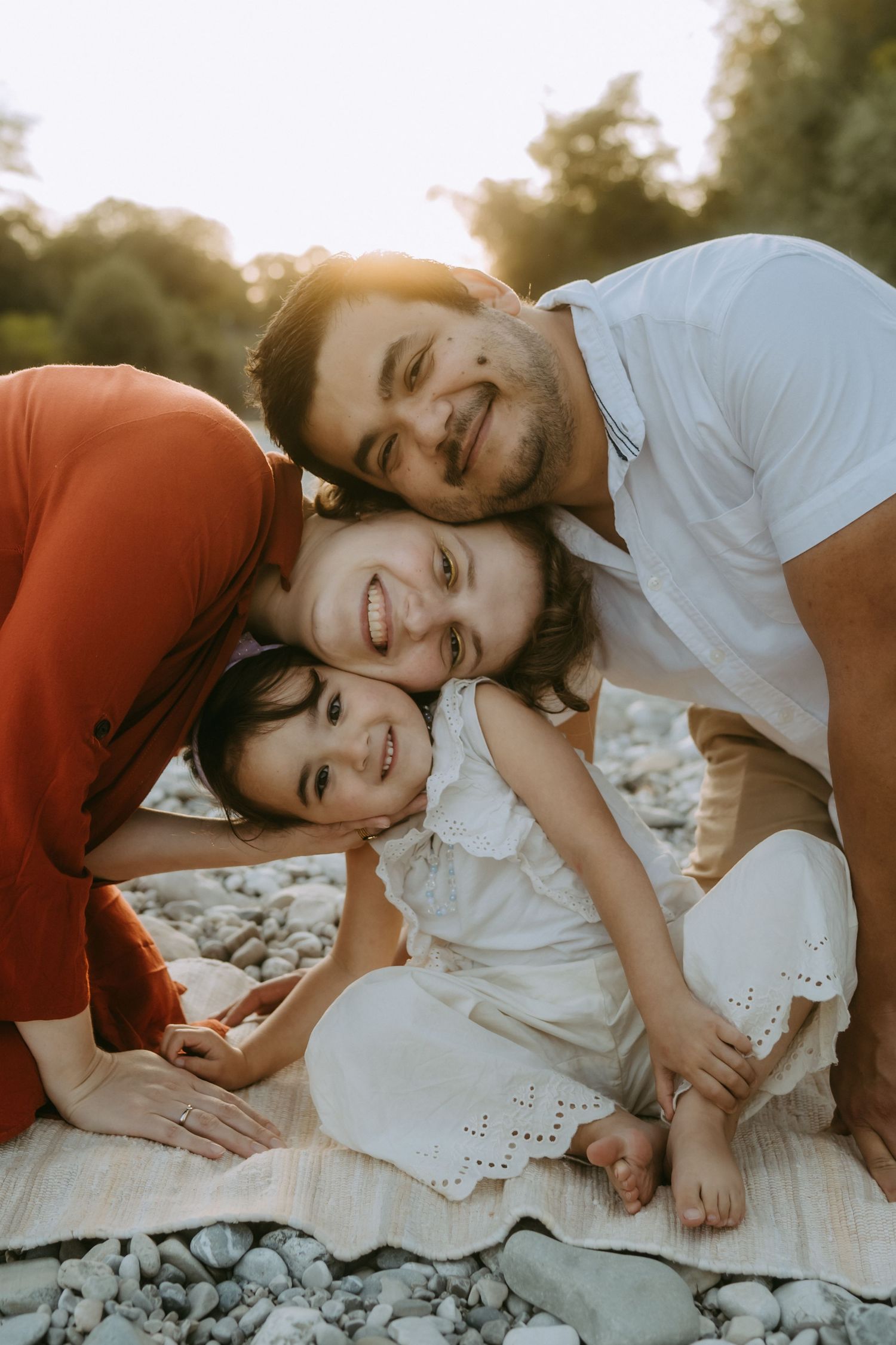 Happy family of three smiling together outdoors on a rocky beach at golden hour sunset.