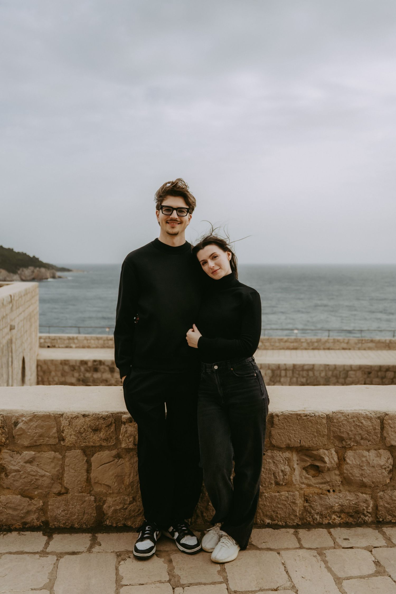 Couple in matching black outfits posing together on a stone wall with ocean and cloudy sky in the background.