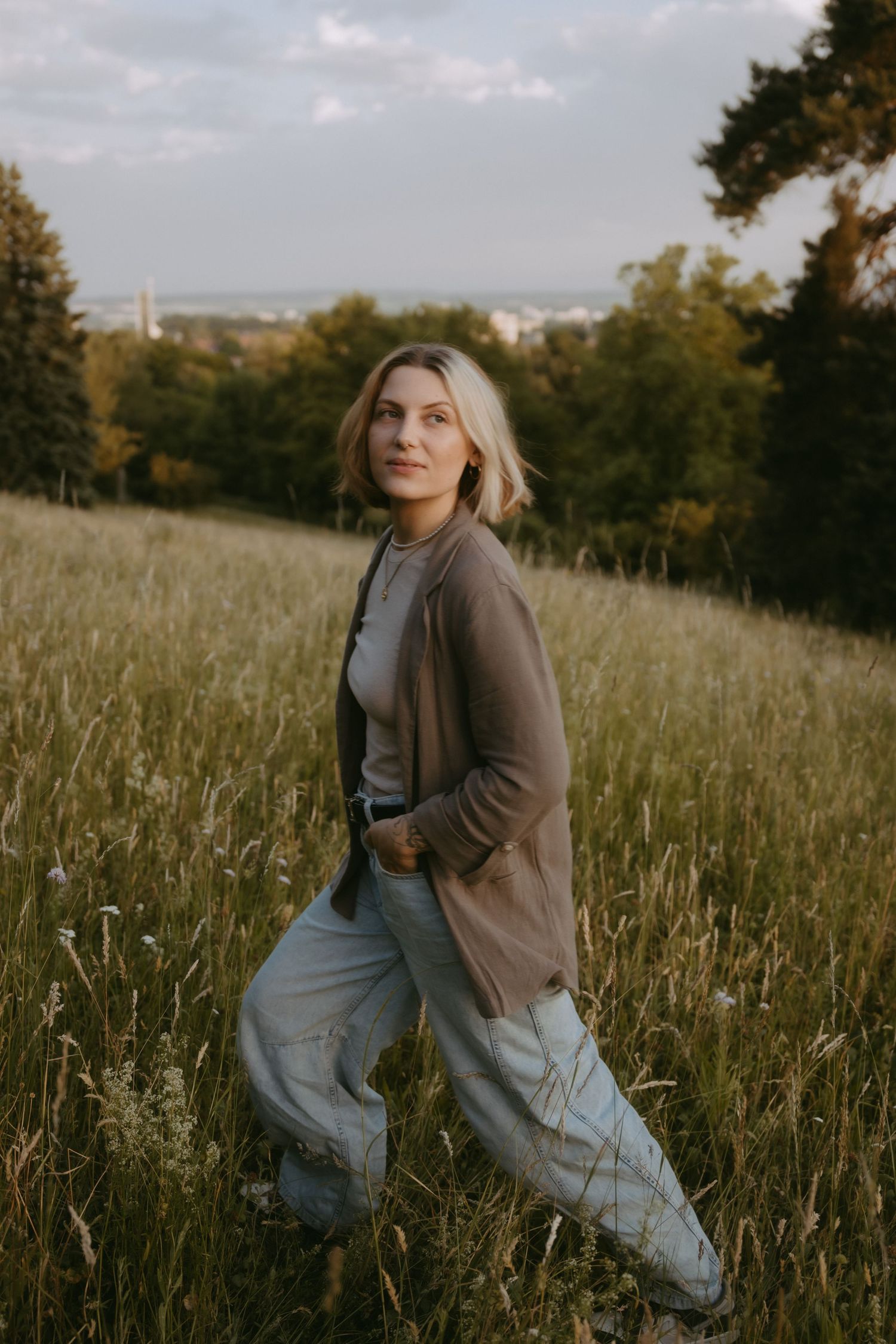 A blonde woman in a brown cardigan and light jeans poses in a lush green meadow with trees and cityscape in background.