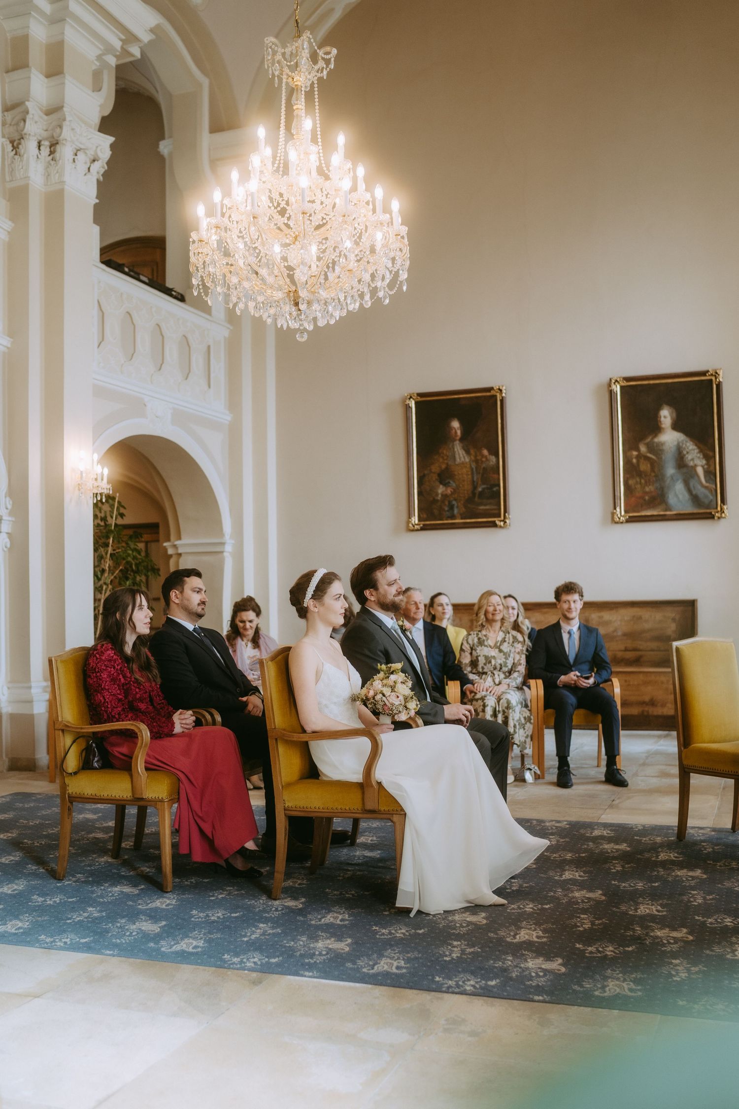 Elegant wedding ceremony in a grand hall with chandelier, bride and groom seated with guests beside portrait paintings.
