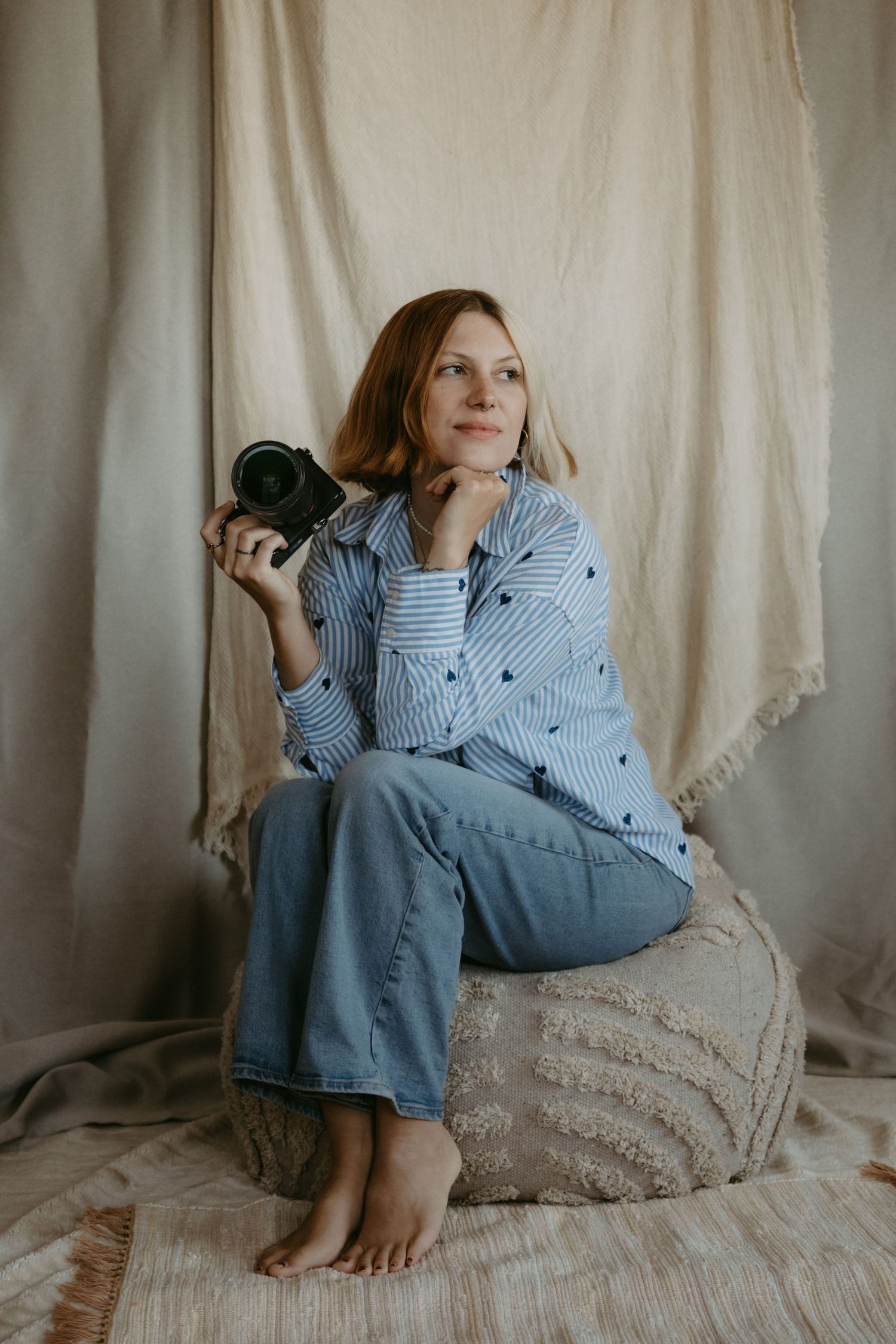 Woman photographer sitting barefoot on ottoman holding camera, wearing striped shirt and jeans against neutral fabric backdrop.