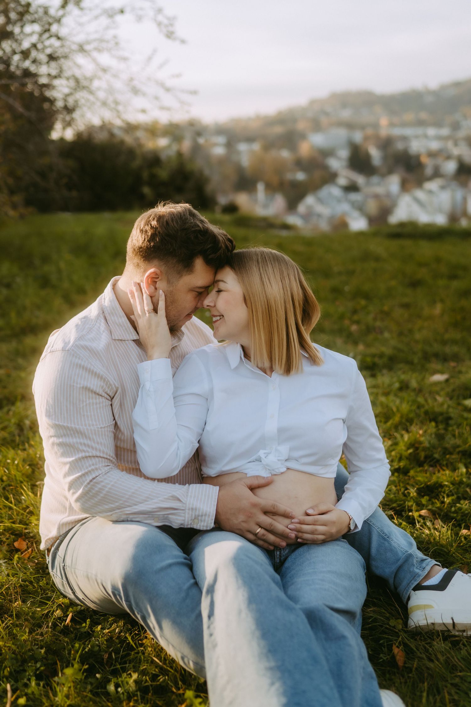 Couple sitting on grass at sunset, man embracing pregnant woman, both smiling at each other with town in background.