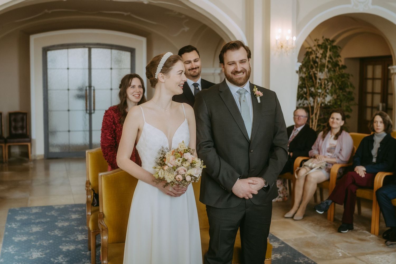 Bride and groom stand at wedding ceremony altar, bride holding bouquet, guests seated in elegant venue.