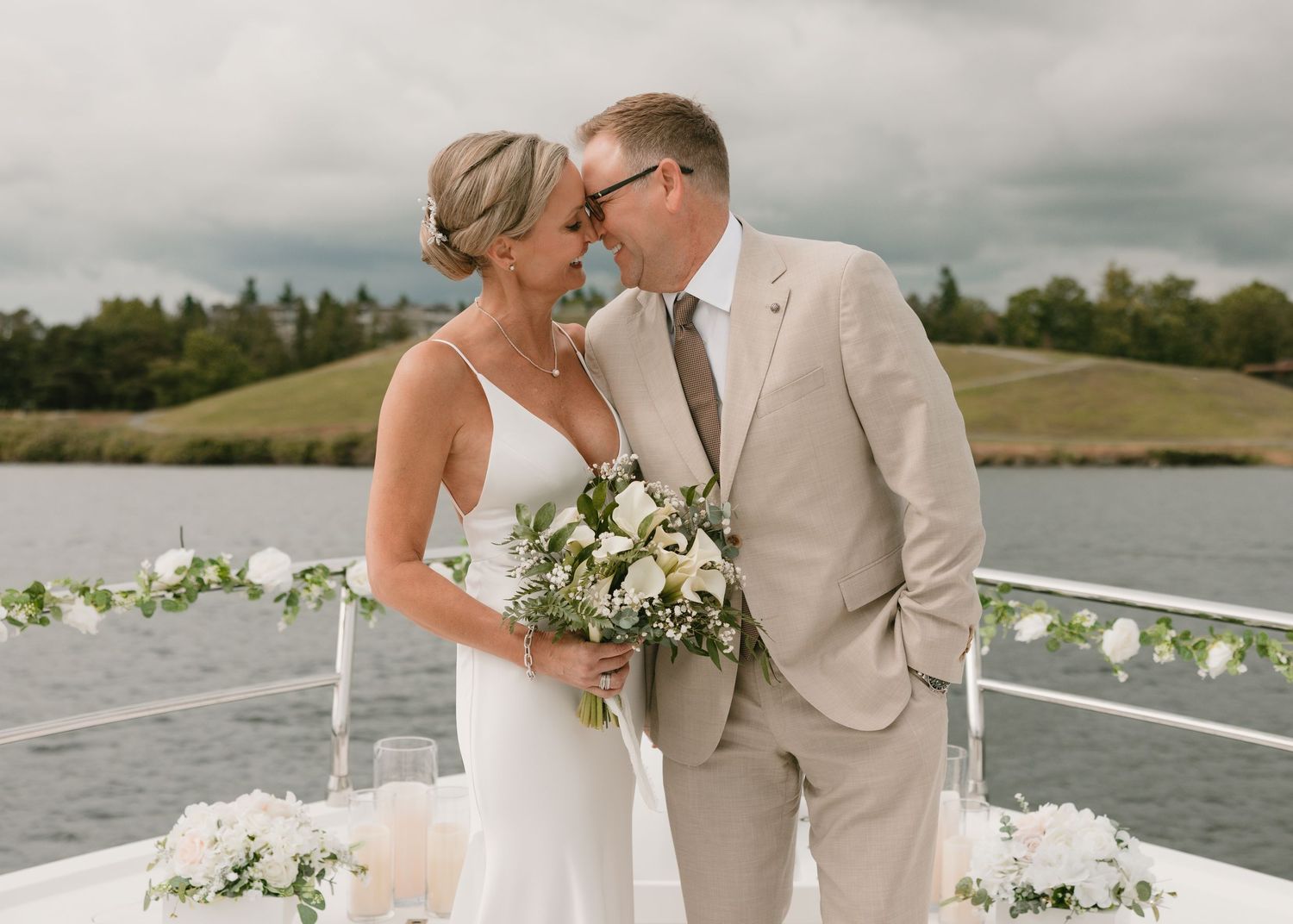 A couple shares an intimate moment on a boat deck decorated with white flowers and greenery by a scenic waterfront.