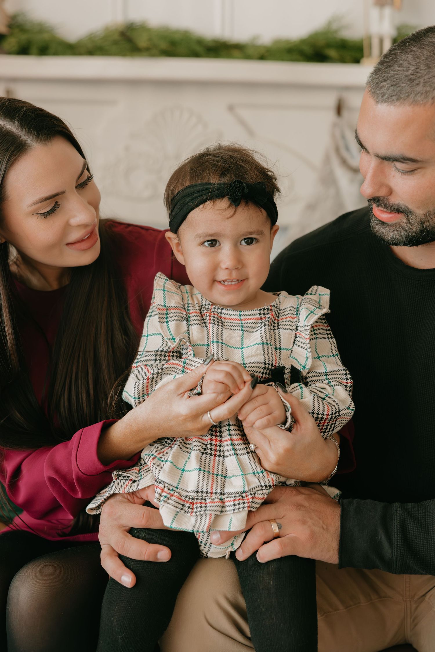 Family sits together on couch with toddler wearing plaid dress during intimate indoor portrait session.