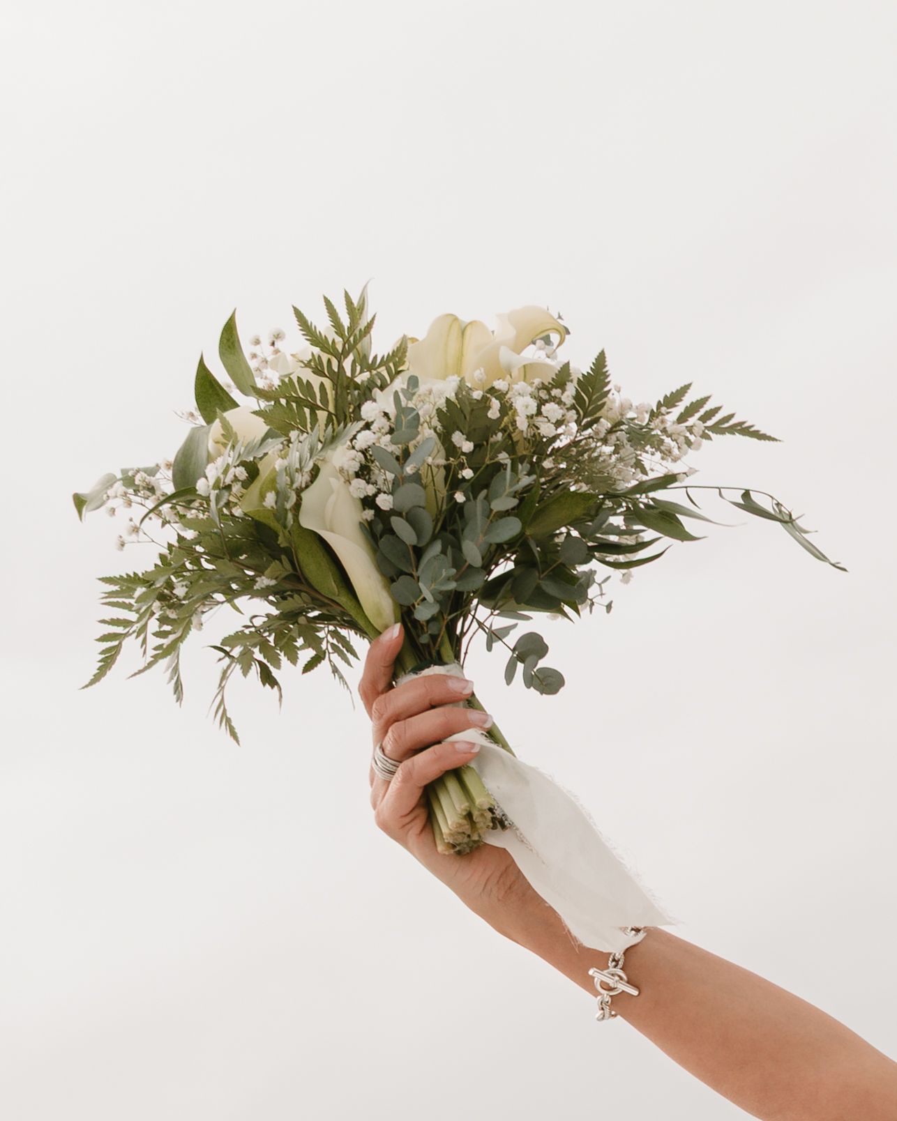 A hand holding an elegant white and green floral bouquet with eucalyptus leaves against a light background.