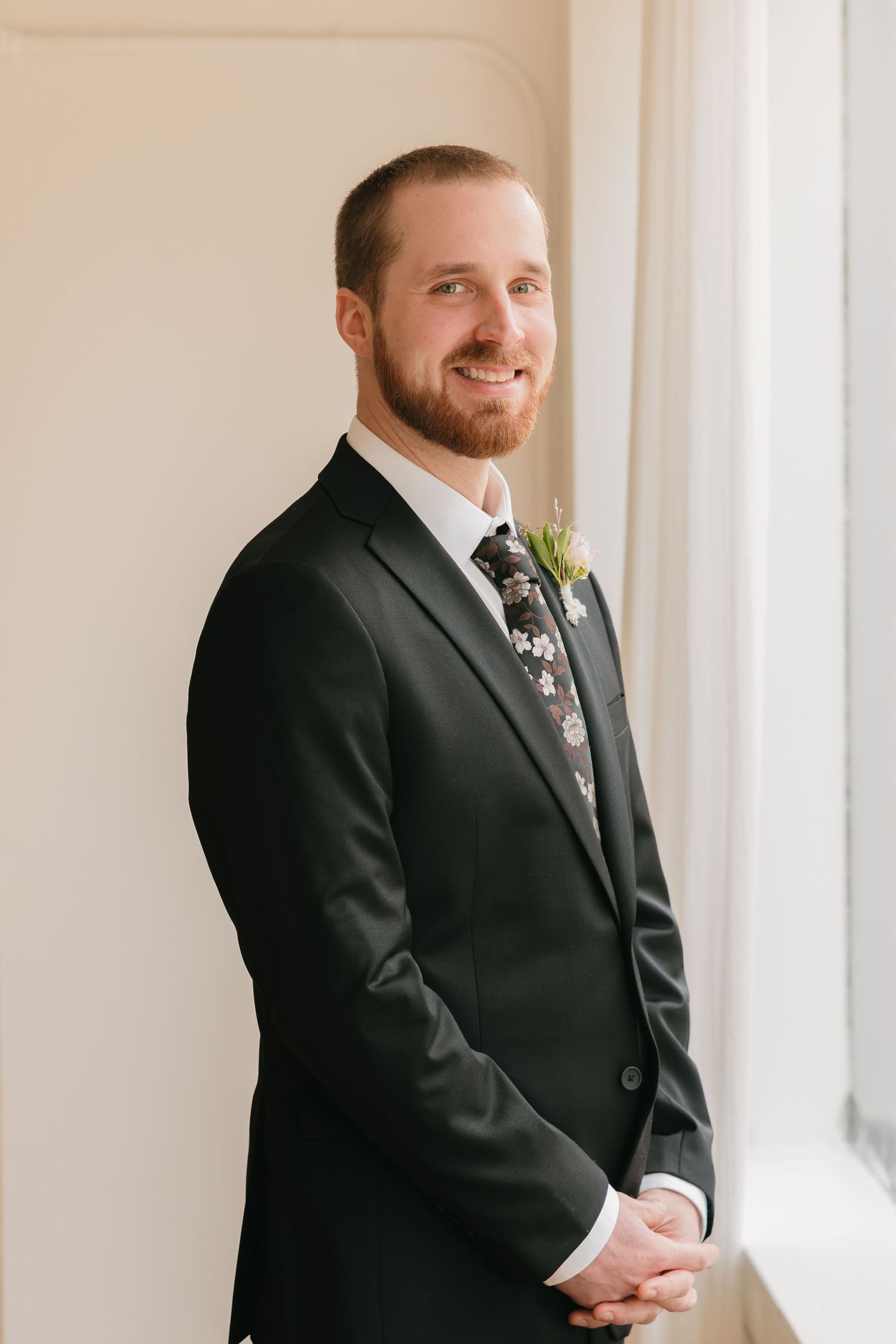 A groom in a black suit and tie stands by a bright window in an elegant indoor setting.
