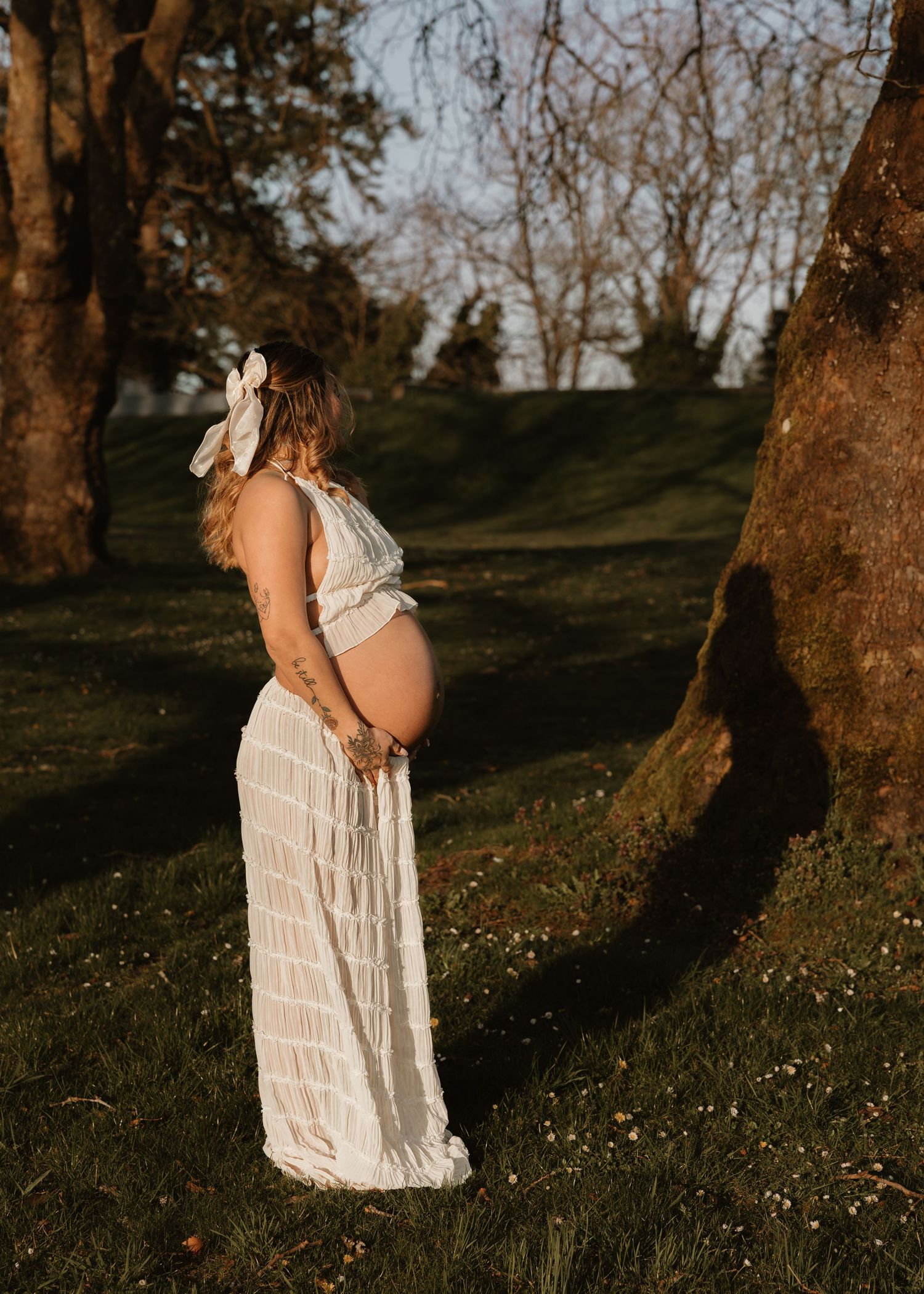 A figure in a flowing white lace dress stands among trees at sunset in a dreamy outdoor setting.