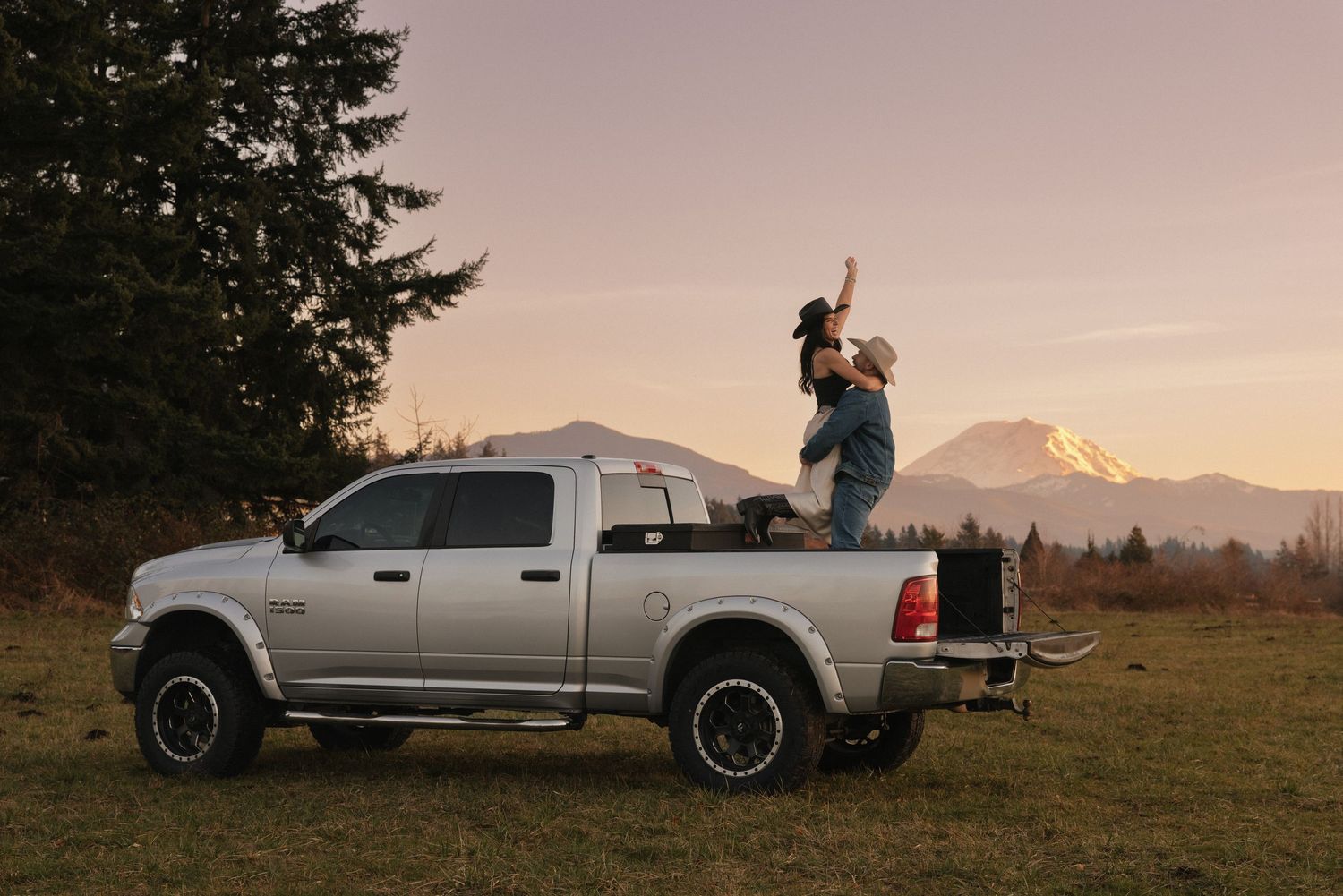 Couple celebrating on a silver Ram pickup truck bed with Mount Rainier at sunset in a scenic field.