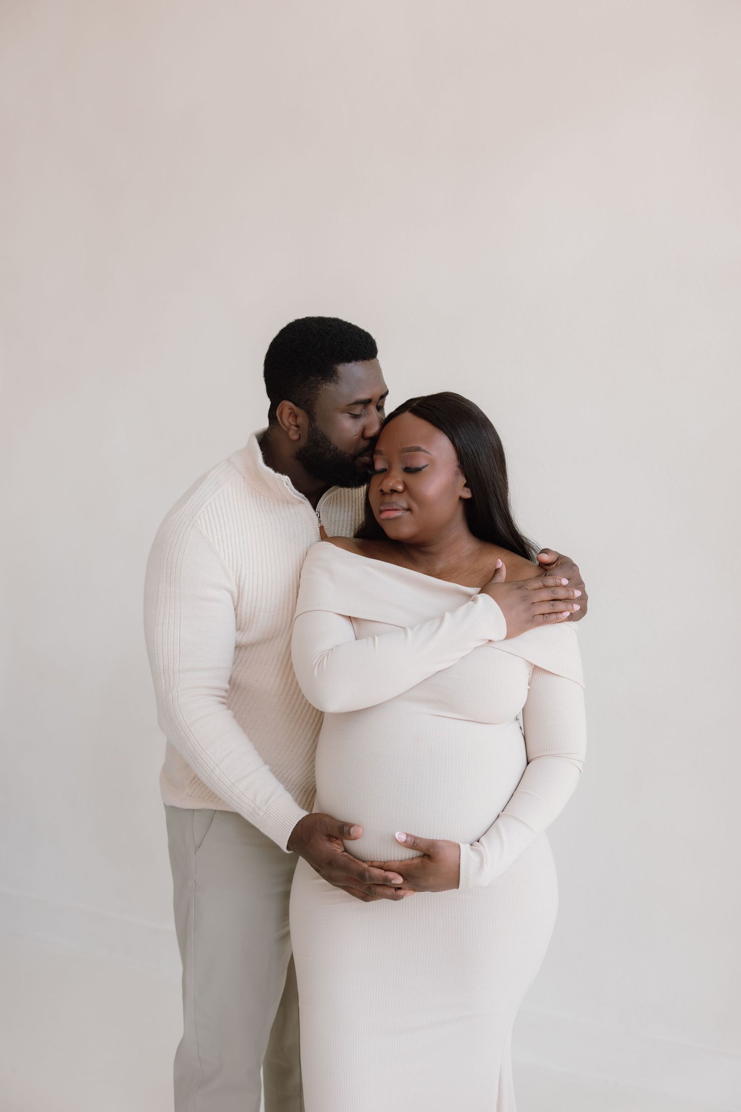 A man kisses a pregnant woman on the cheek as she cradles her baby bump, both dressed in white.