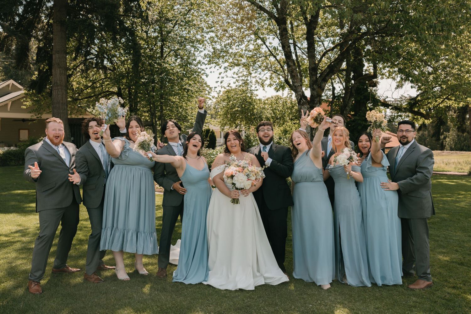 Wedding party in light blue dresses and dark suits poses together on a sunny lawn during outdoor celebration.