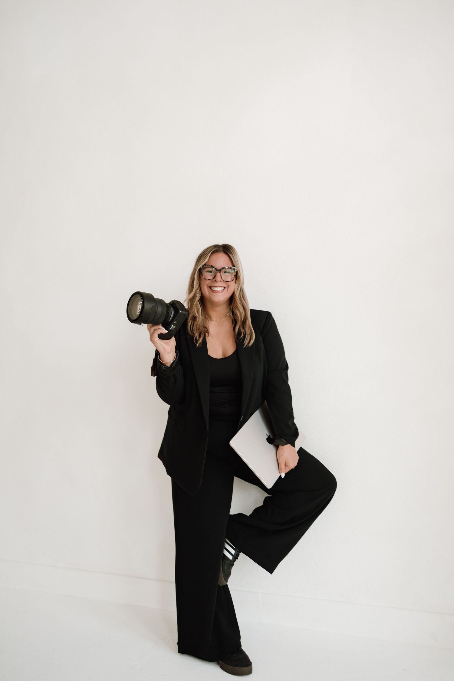 Professional photographer in black attire poses with camera equipment against white studio backdrop.